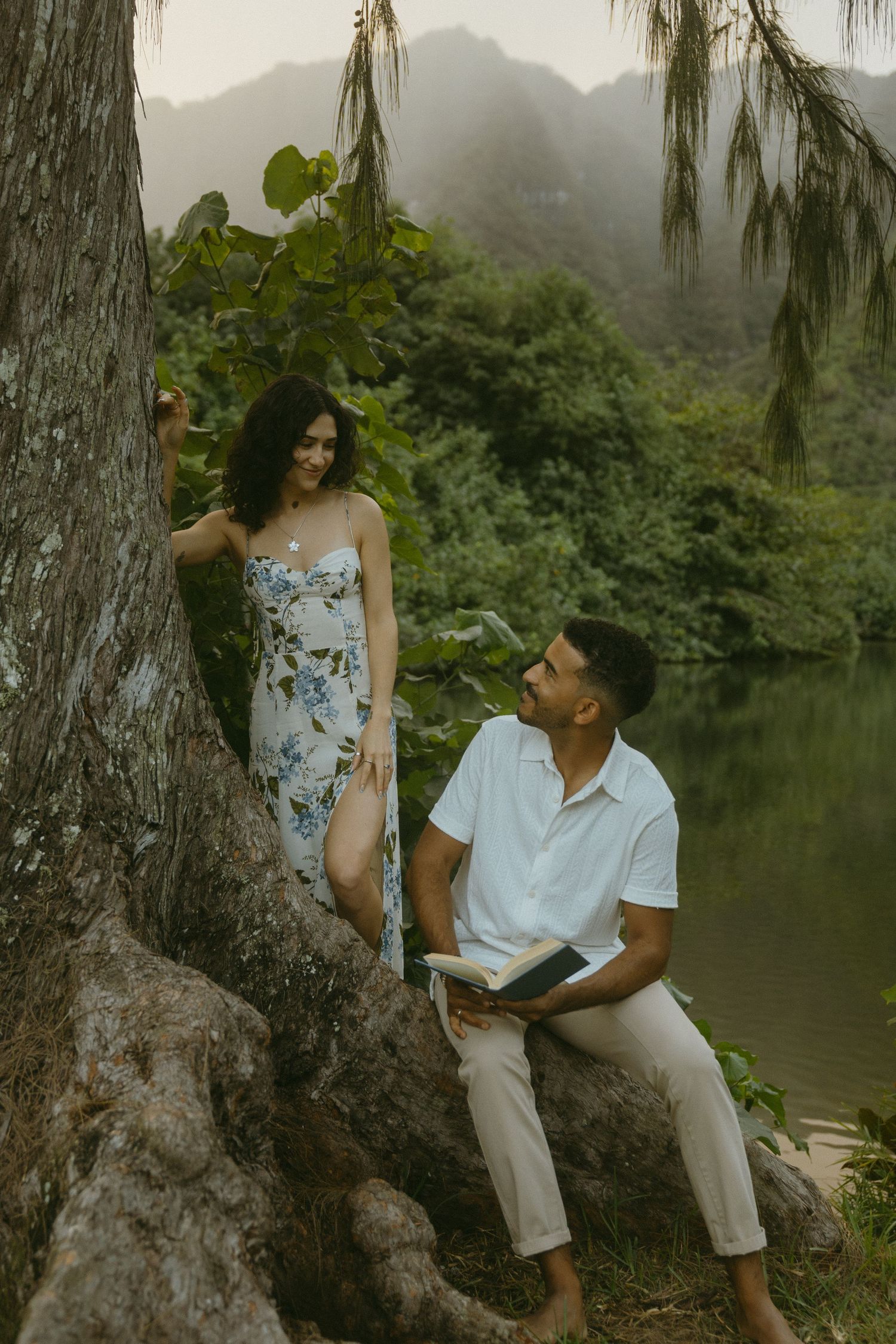 A couple posing together under a willow tree by a serene lake with mountains in the distance.