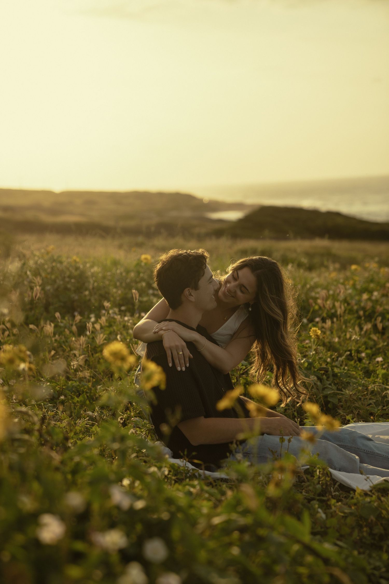 Two people lying in a golden meadow at sunset surrounded by wildflowers and tall grass.