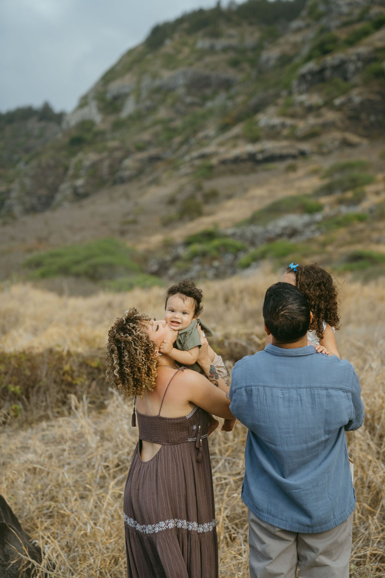 A couple stands together taking photos against a scenic hillside backdrop in casual attire.