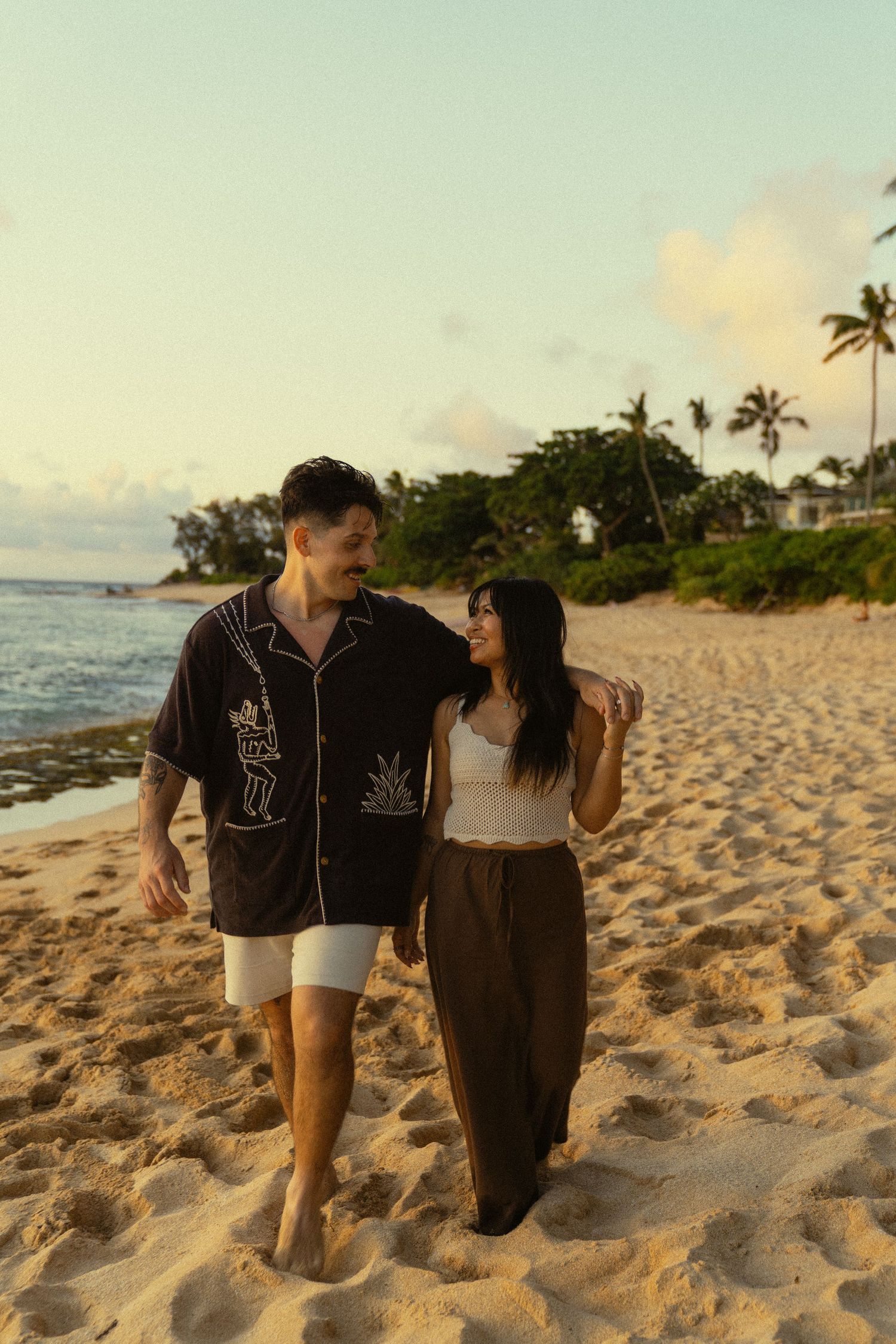 A couple walks together on a sandy beach at sunset wearing matching dark outfits.