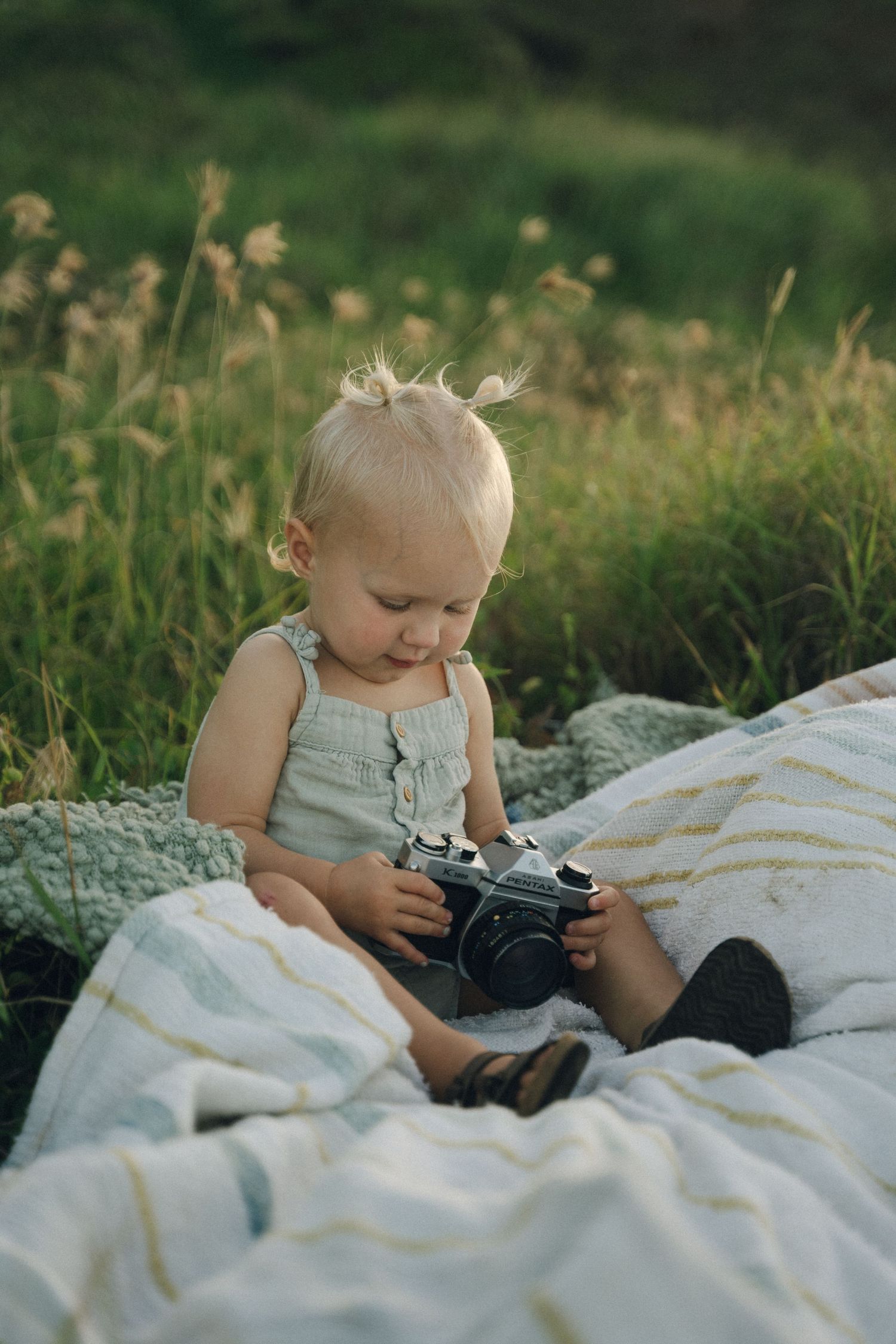 Small child exploring a vintage camera while sitting on a blanket outdoors.