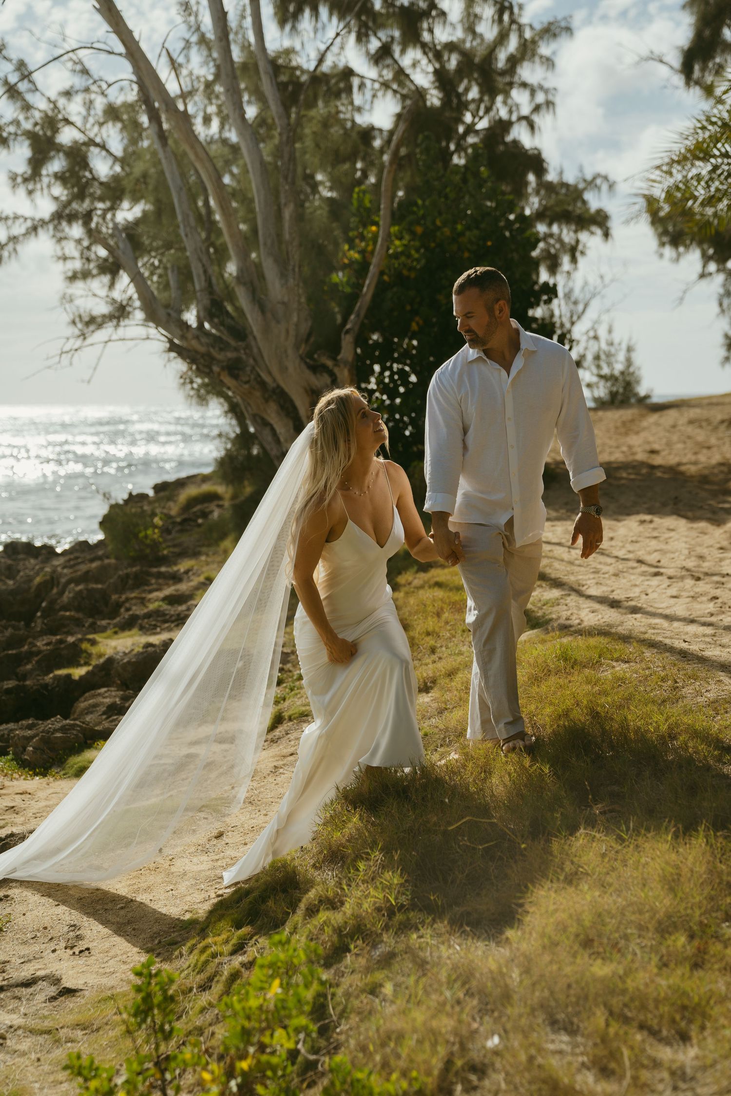 Couple in white attire walks along scenic coastal path with flowing veil in breeze.