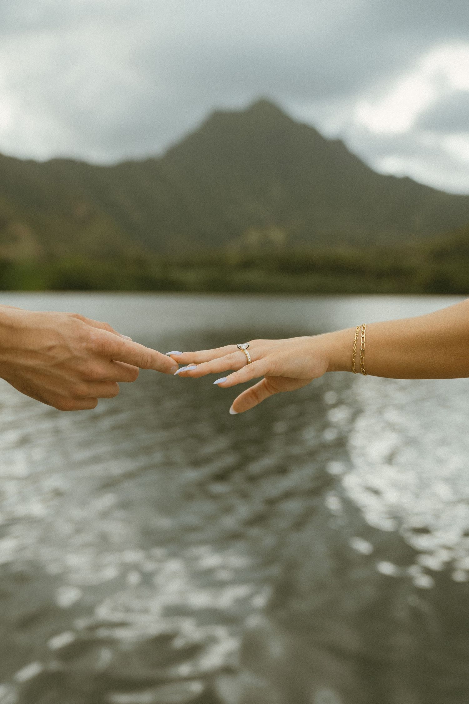 Hands reaching across water with mountains and cloudy sky in the background.
