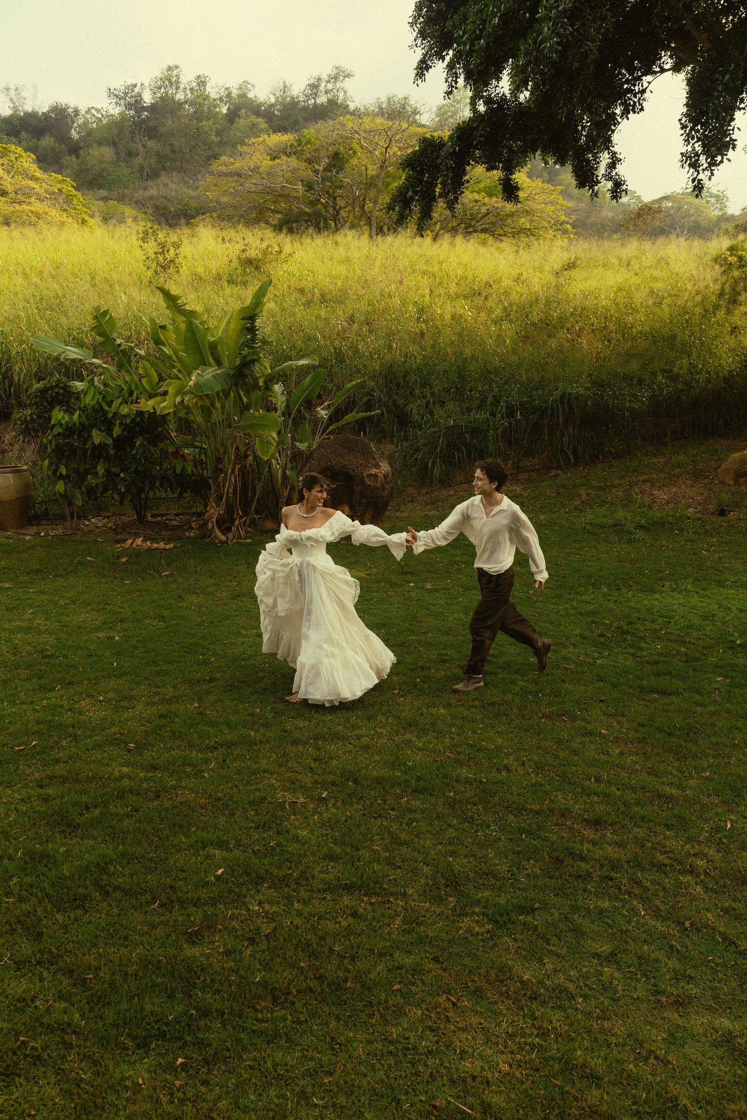 Two figures dance playfully across a grassy field in vintage-style attire at golden hour.