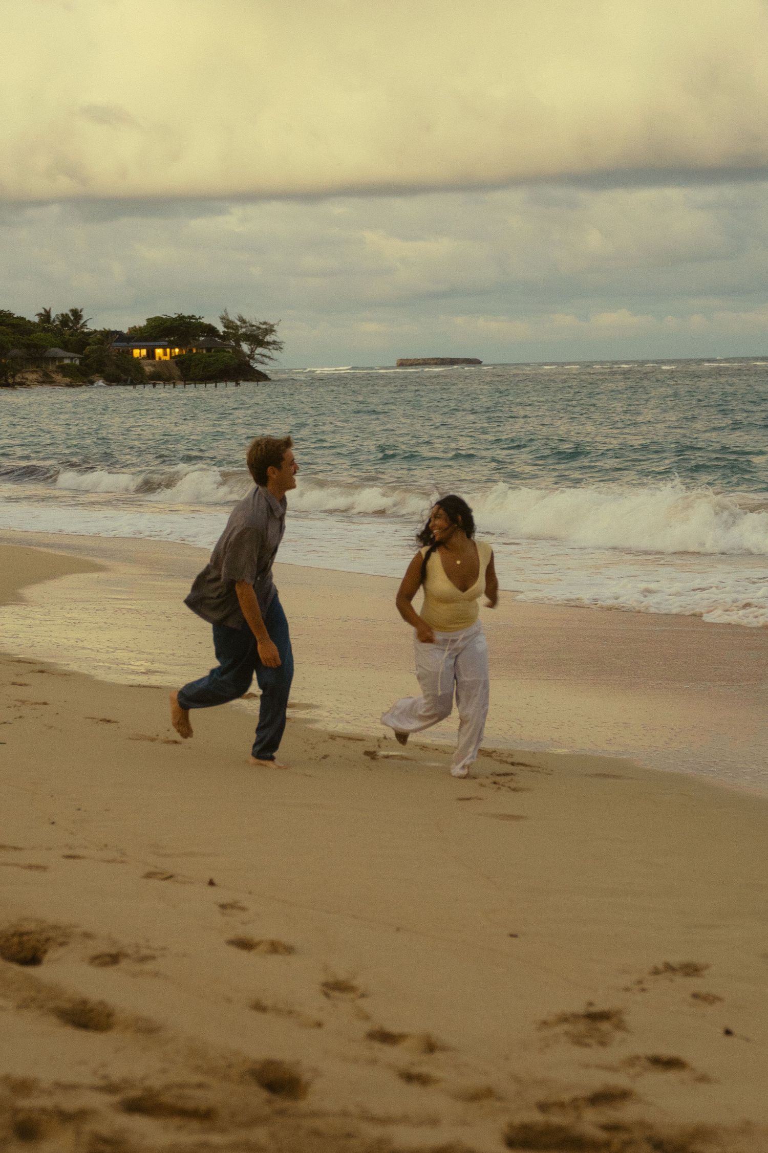 Two people chase each other playfully across a sandy beach at sunset.