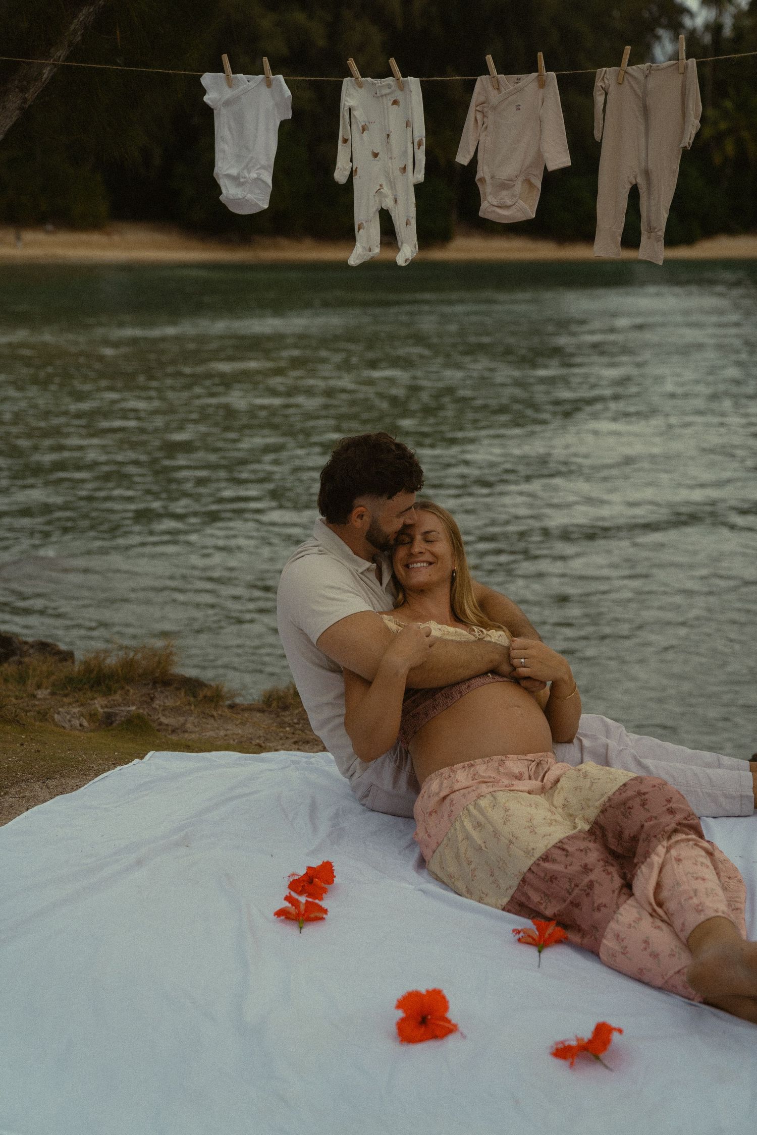 A couple shares an intimate moment on a white blanket with orange flowers by the water.