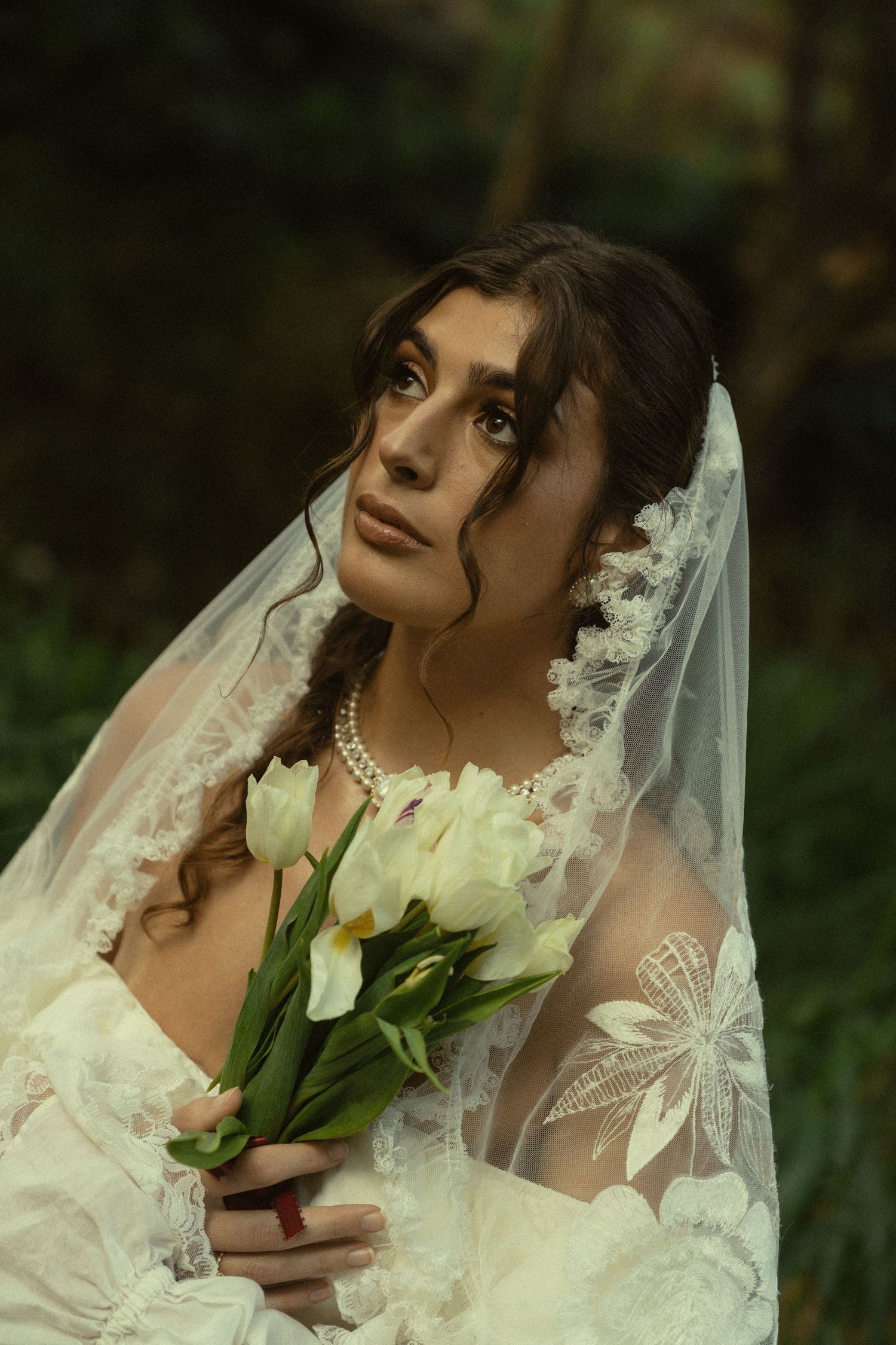 A bride in a lace veil holds white tulips while posing against a dark forest background.