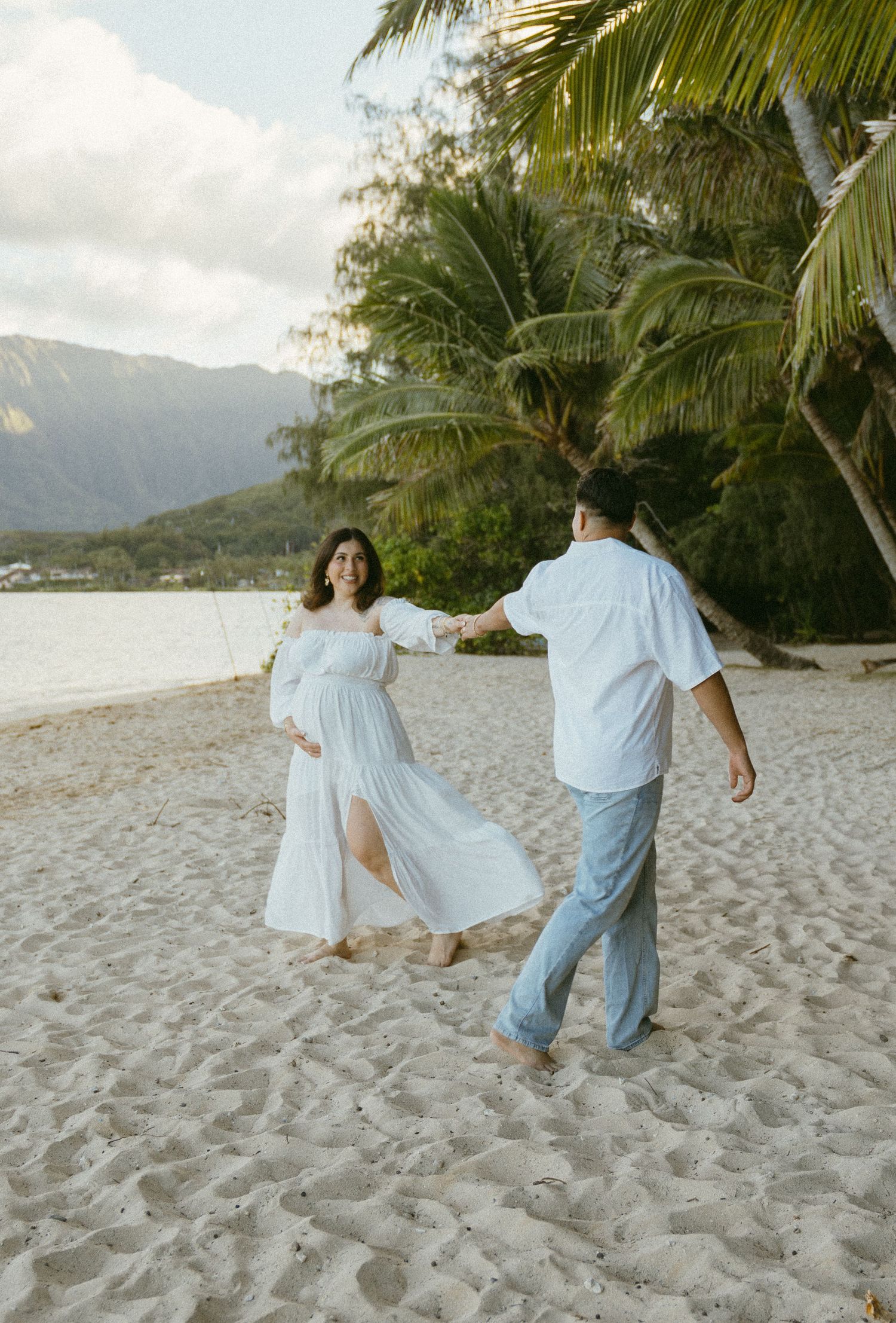 Two people in light clothing dance joyfully on a sandy beach with palm trees and mountains.