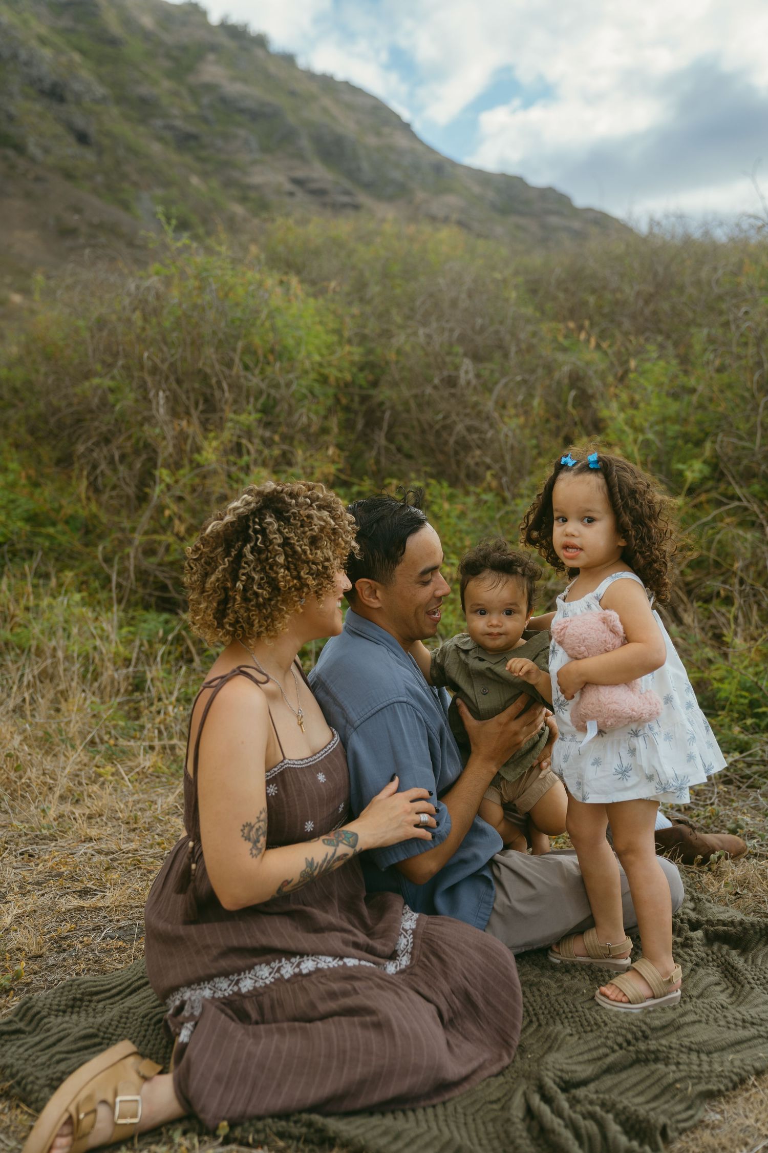 Family shares intimate moments while sitting on a green blanket against mountainous landscape.