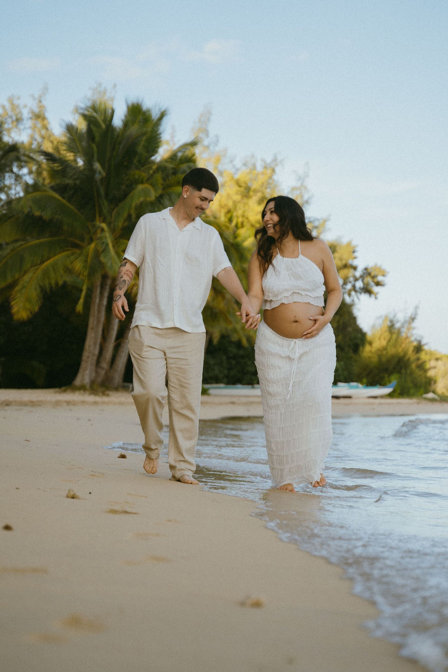 A couple walks along a tropical beach shoreline in matching white and beige outfits.