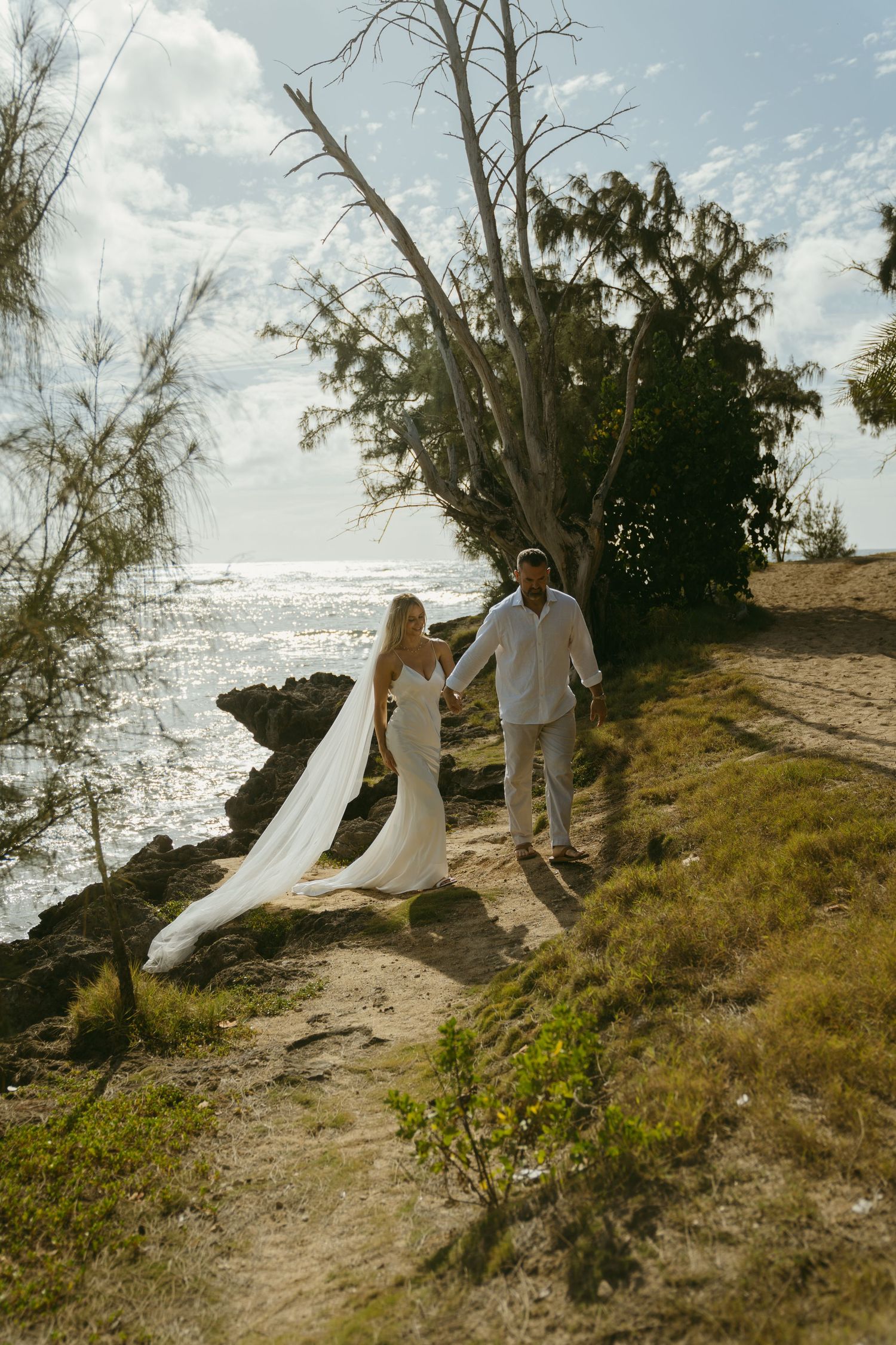 A couple in white attire poses on a scenic coastal cliff overlooking the ocean.