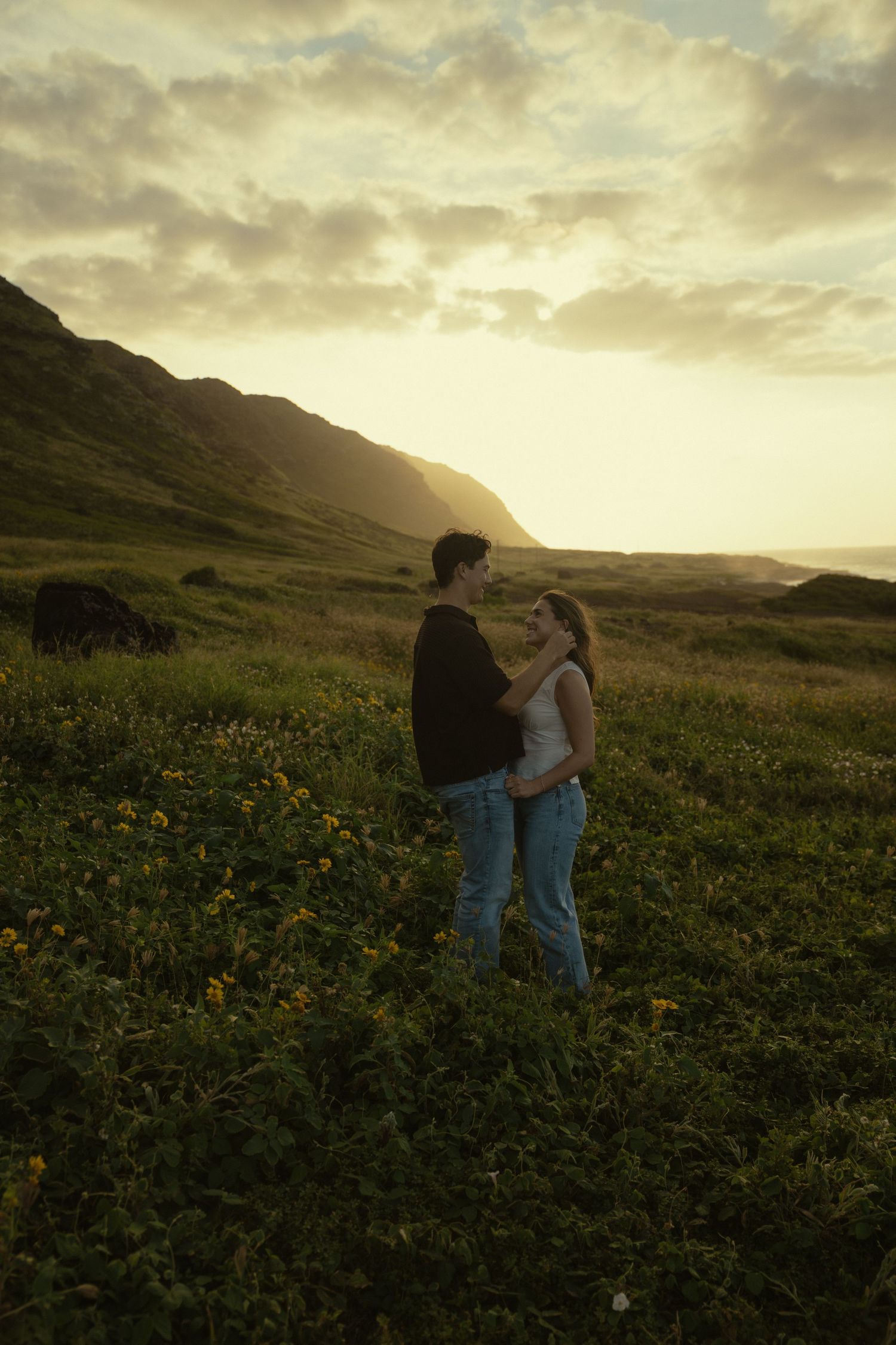 Two people embrace in a golden mountain meadow at sunset while surrounded by wildflowers.