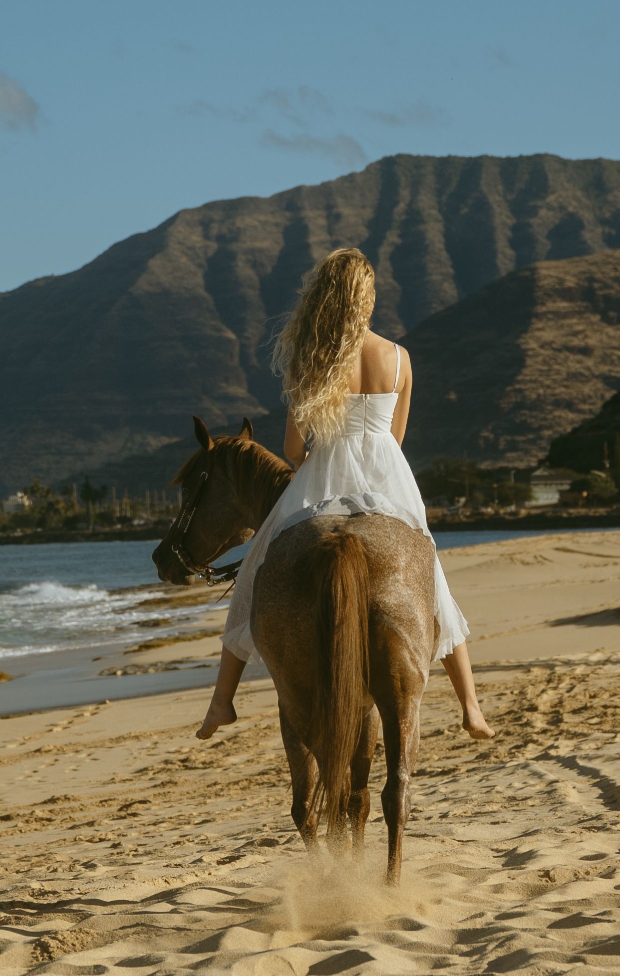 A white-dressed figure rides horseback on a sandy beach with dramatic mountain peaks in the distance.