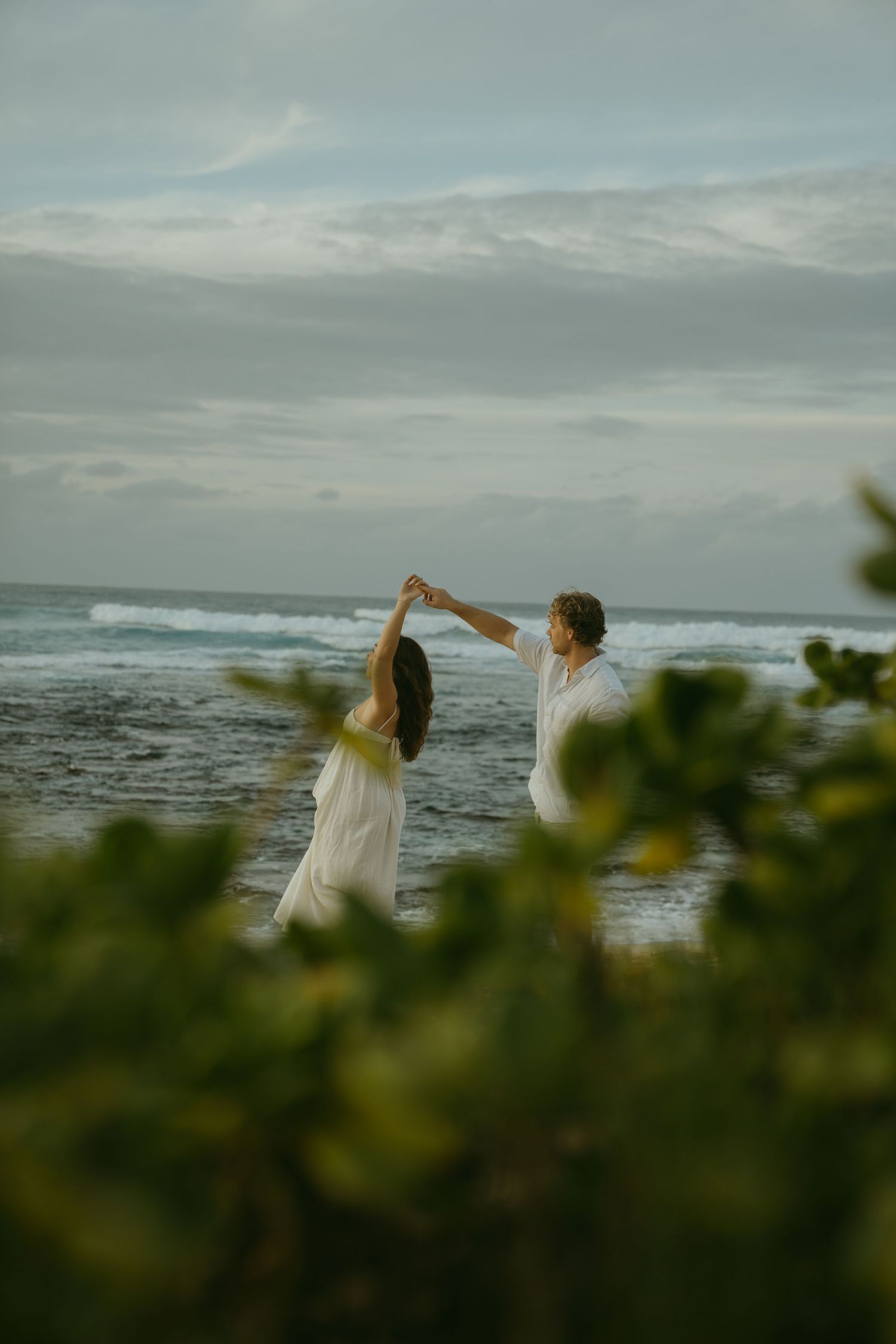 Two people dancing together on a beach with ocean waves and foliage in the foreground.