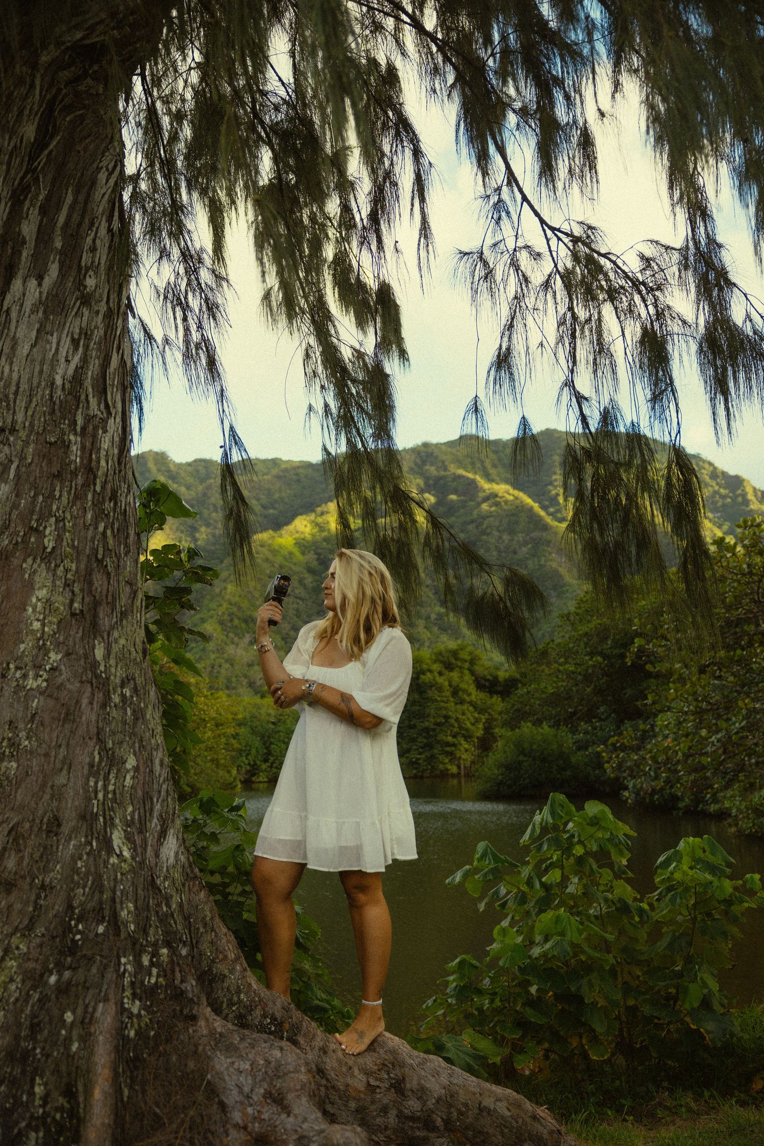 Woman in white dress standing on tree root against scenic mountain backdrop.