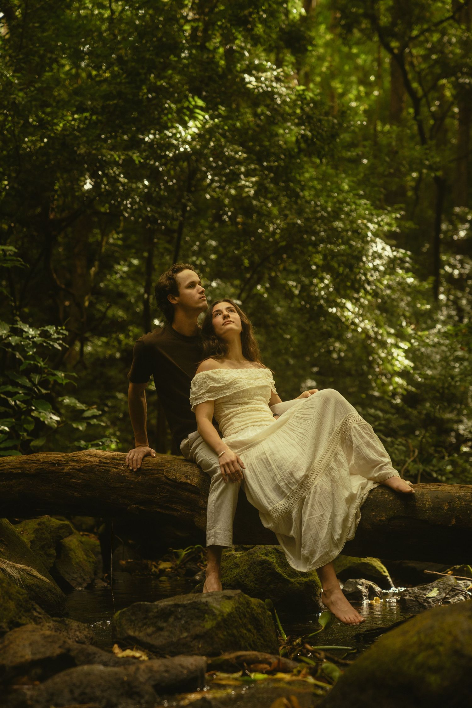 A couple shares an intimate moment while sitting on rocks in a dreamy forest setting.