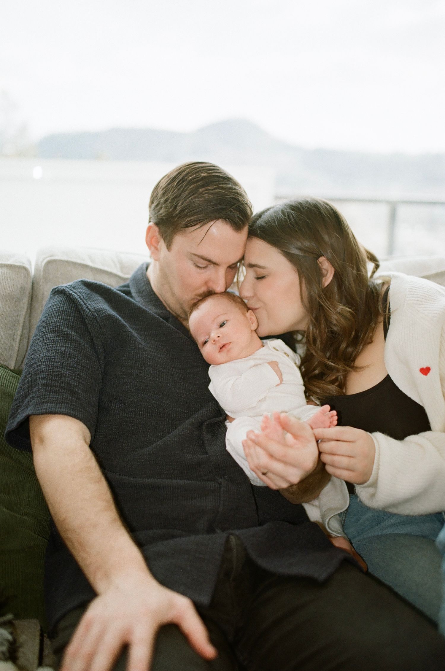 Tender family moment on a cozy grey couch with mountains visible through window.