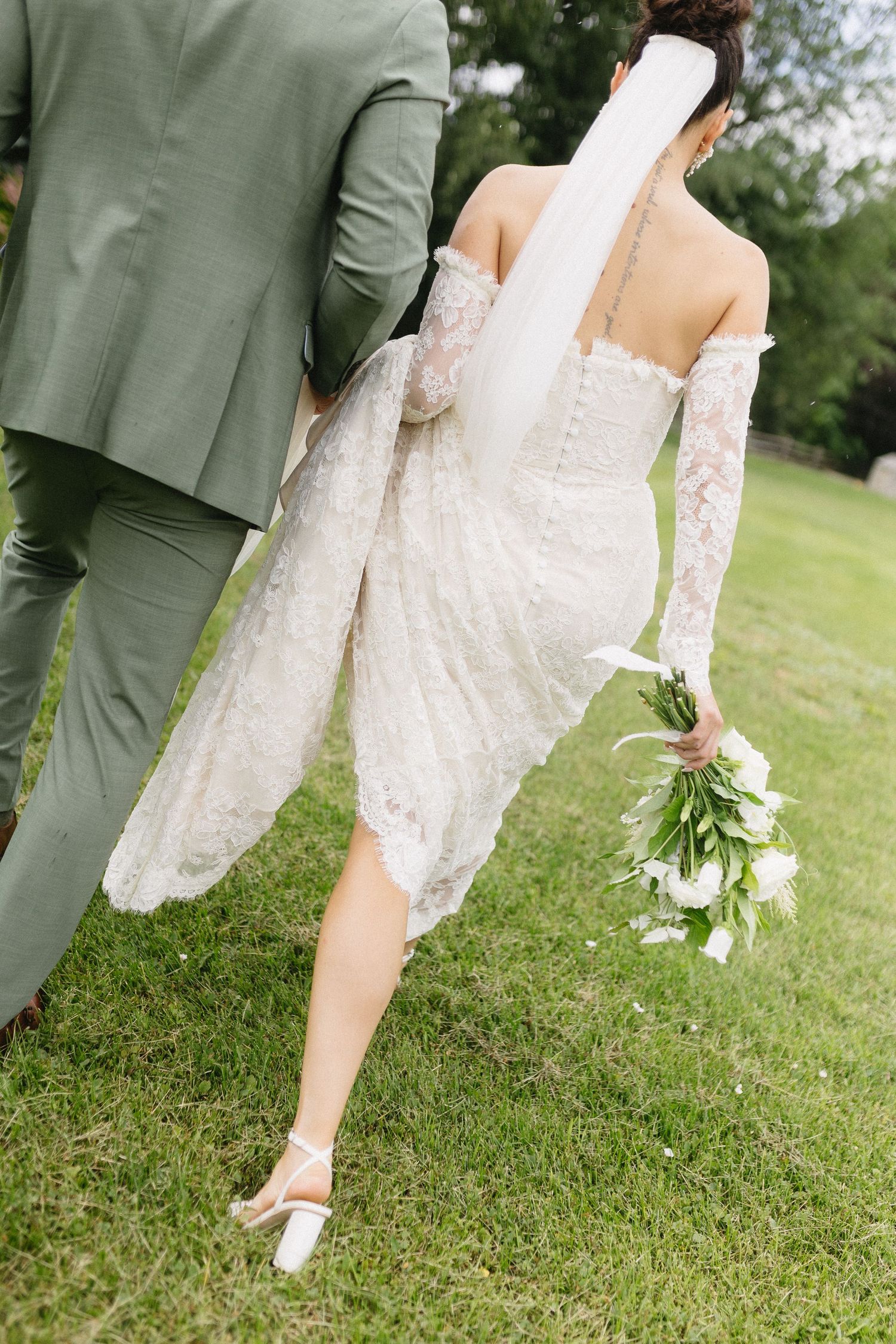 Back view of a couple in sage green suit and lace wedding dress holding a white floral bouquet.