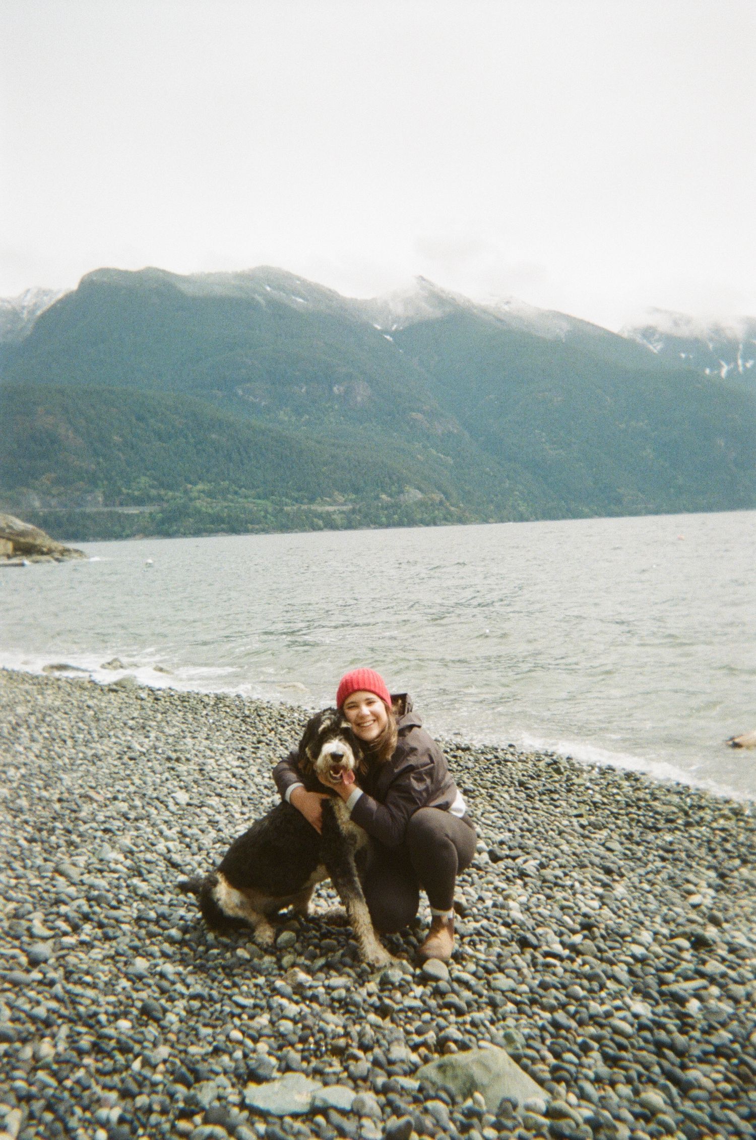 Person in red beanie and winter coat sitting with dog on rocky beach near mountain landscape.