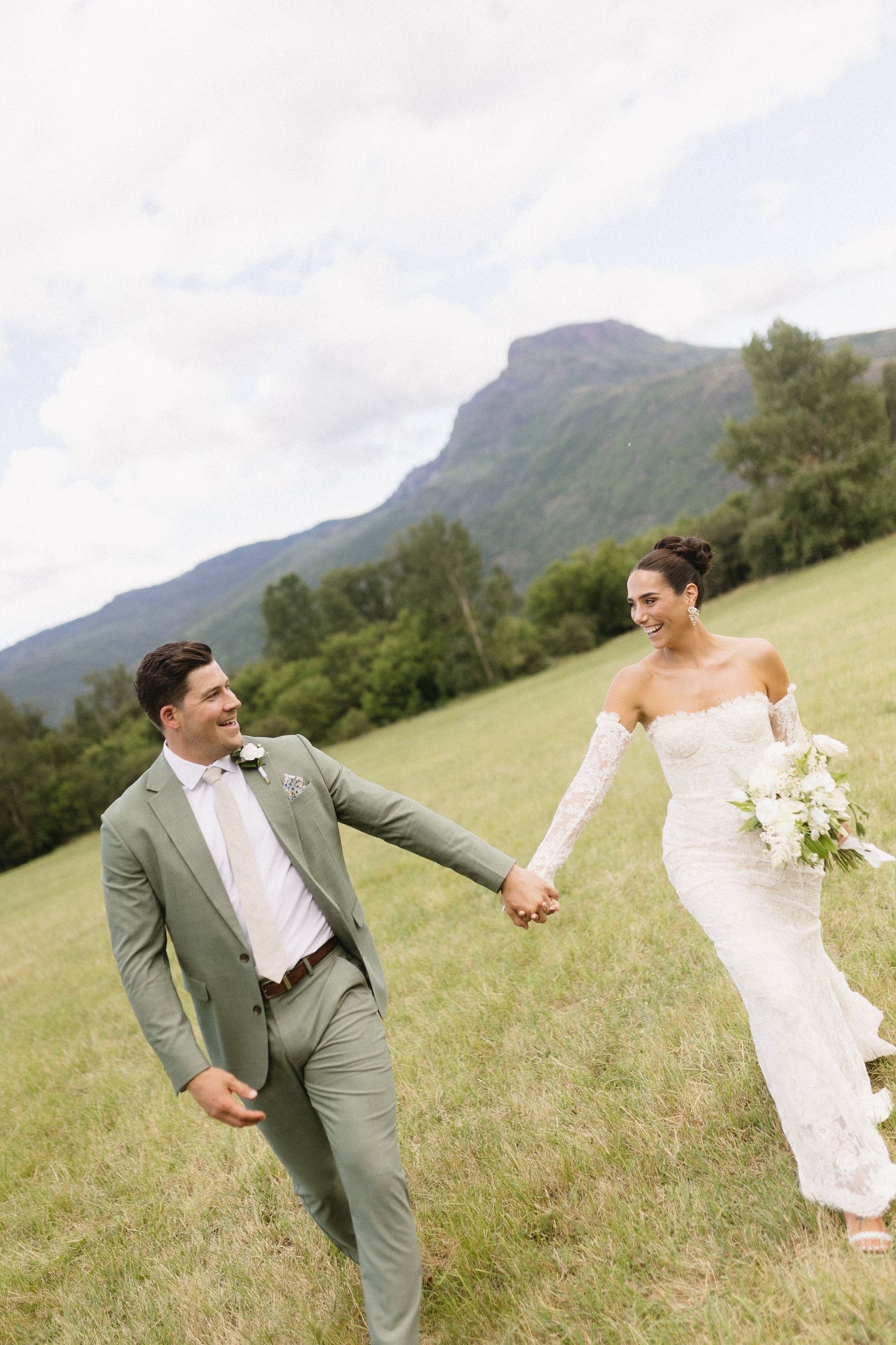 A couple in wedding attire hold hands and walk joyfully across a grassy hillside with majestic mountains in the background.