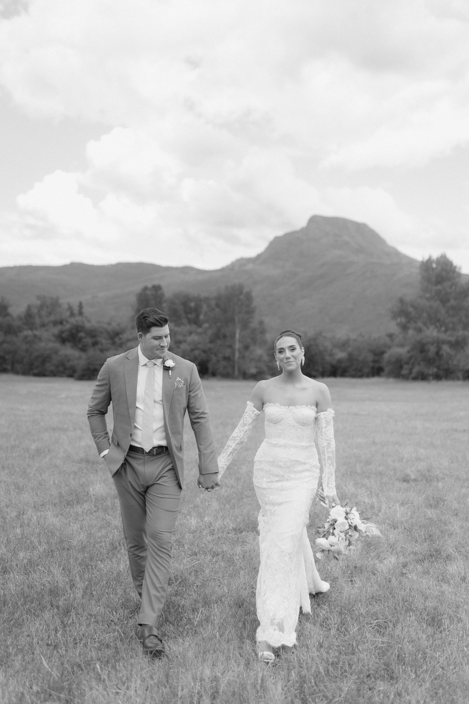 A couple walks hand in hand through a meadow with mountains in the background.