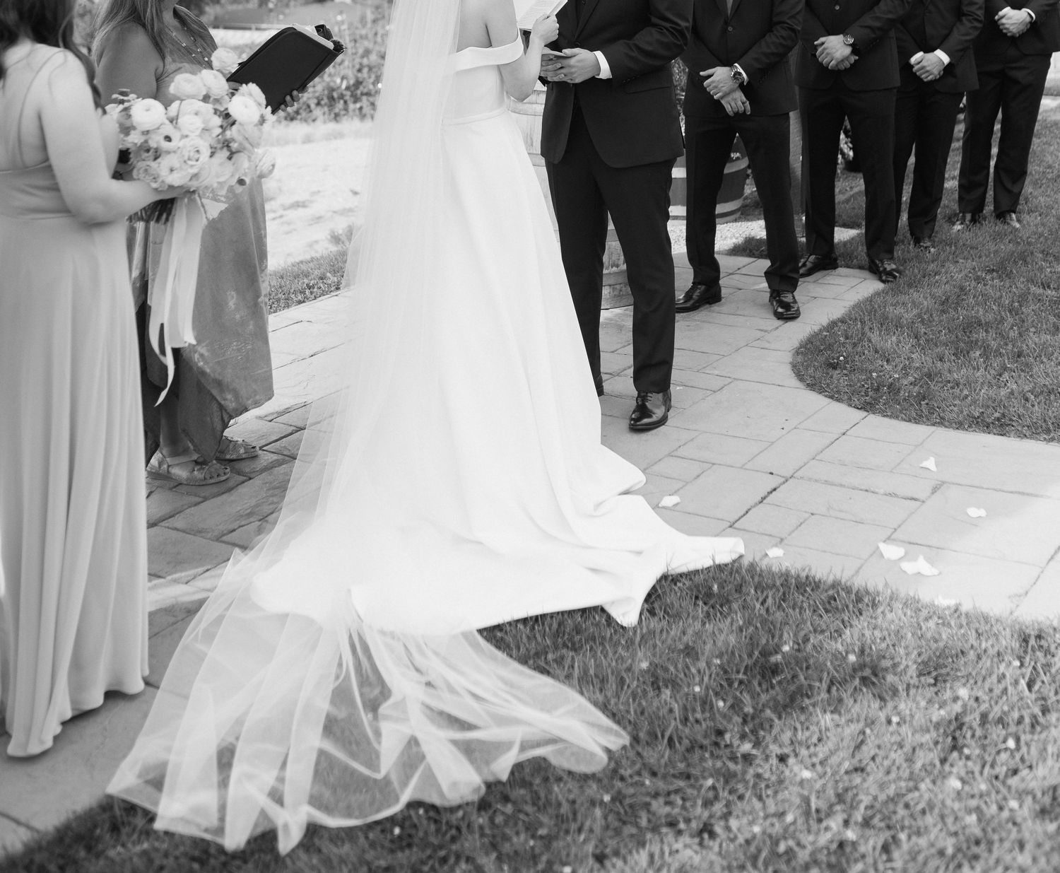 A wedding ceremony party stands in a curved line formation on grass in black and white photography.