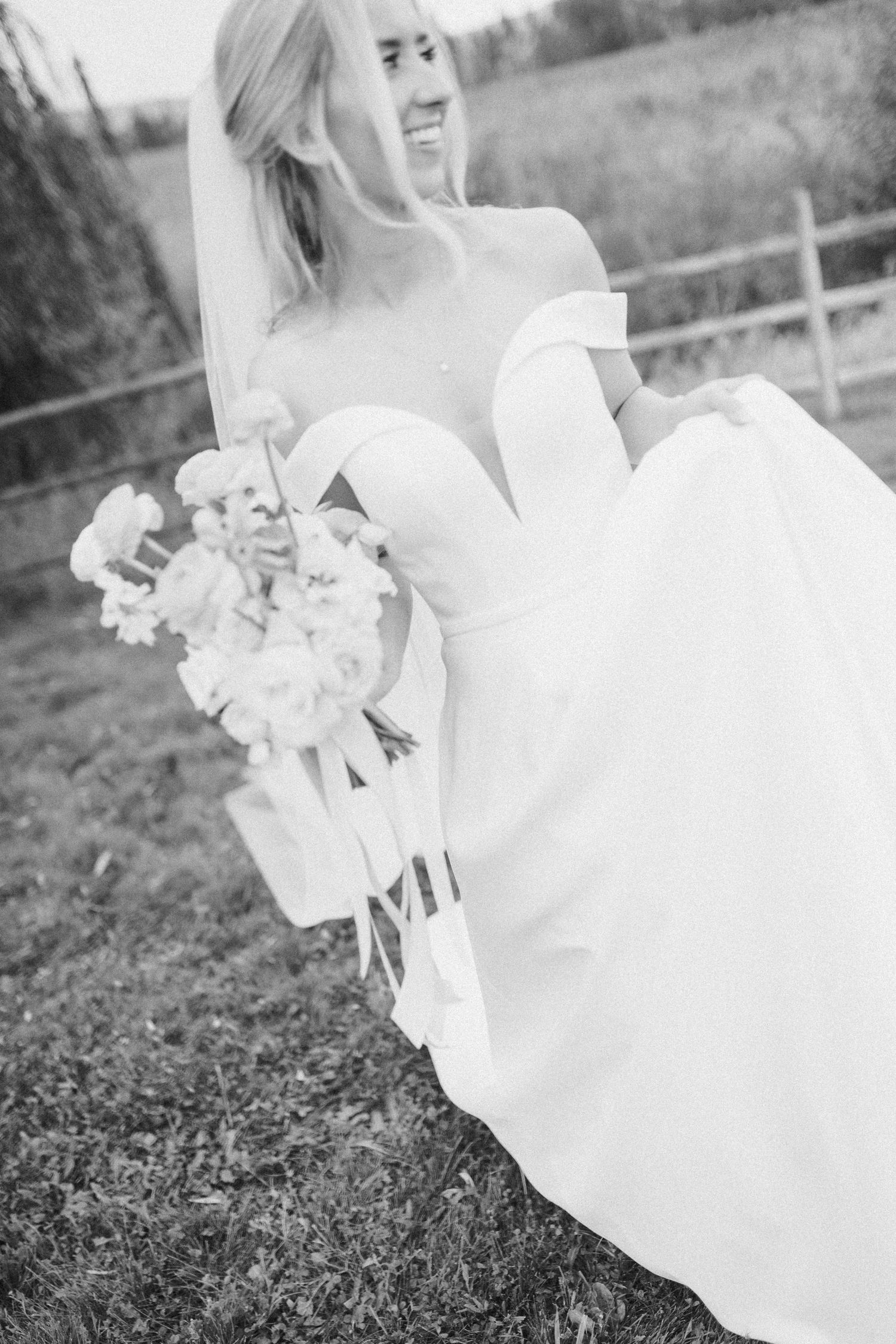 A bride in an elegant off-shoulder white gown holds a bouquet while standing outdoors by a rustic wooden fence.