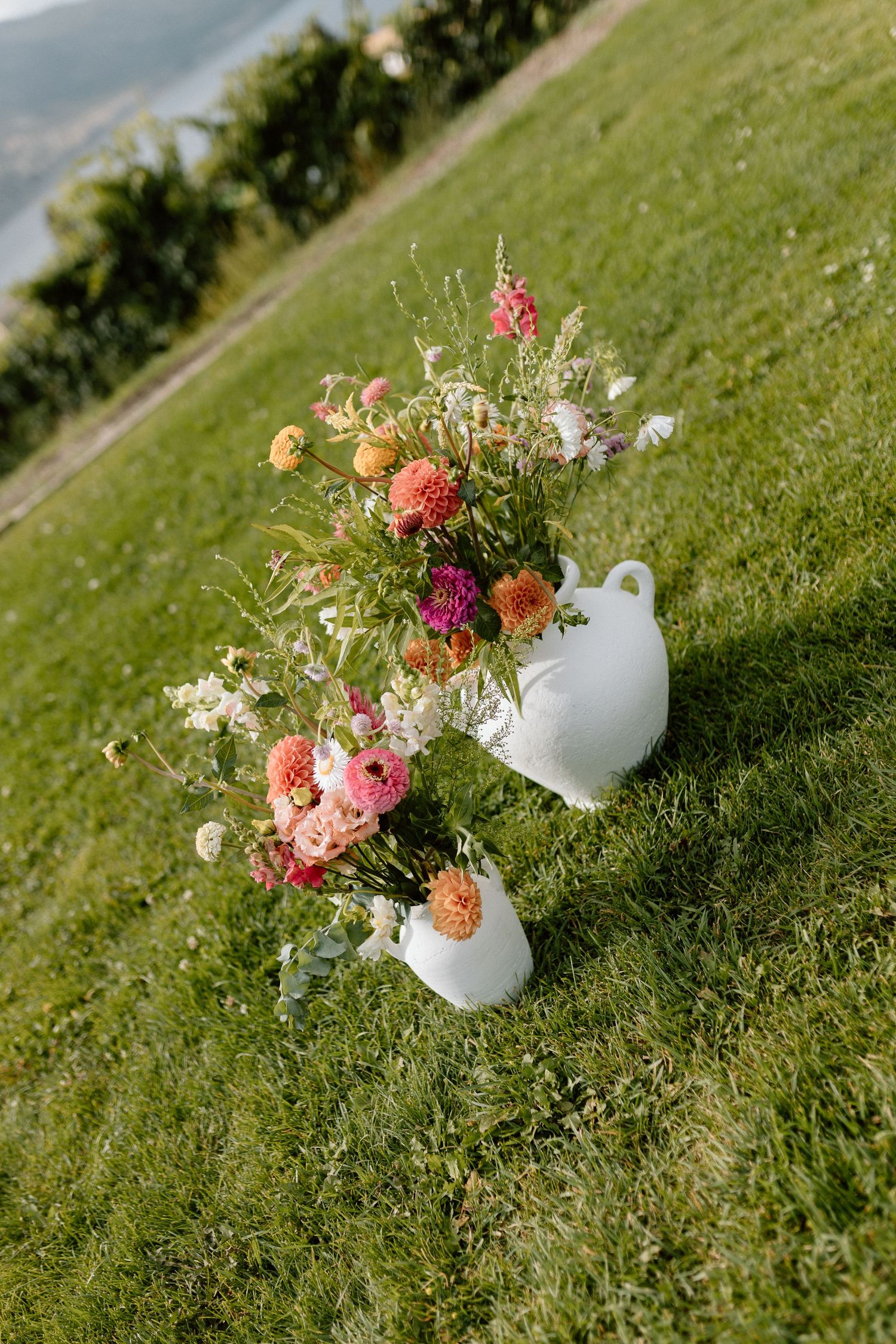 Colorful floral arrangements with pink and peach blooms displayed on grass at an outdoor wedding.