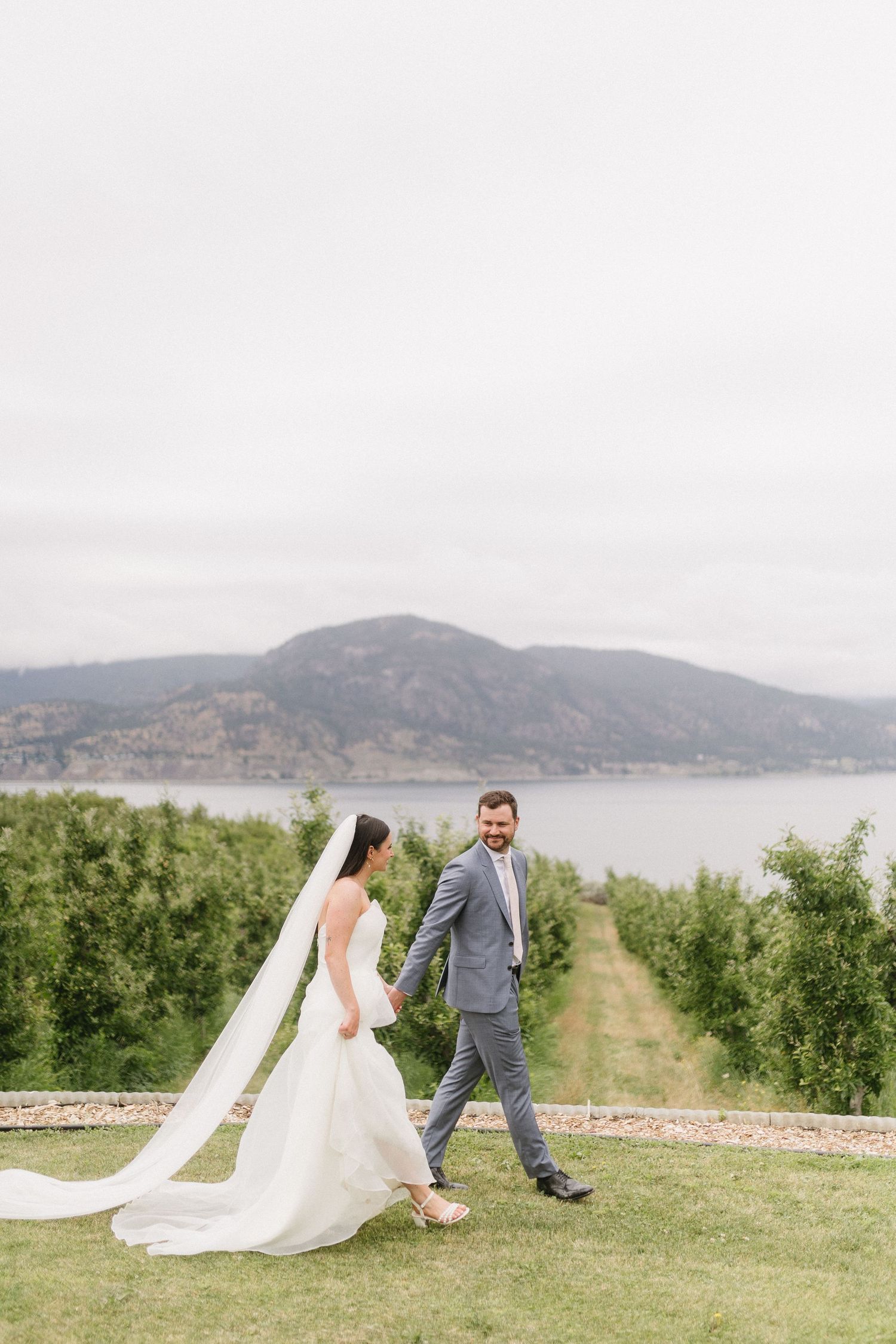 A couple walks hand in hand on a grassy path overlooking a scenic mountain lake.