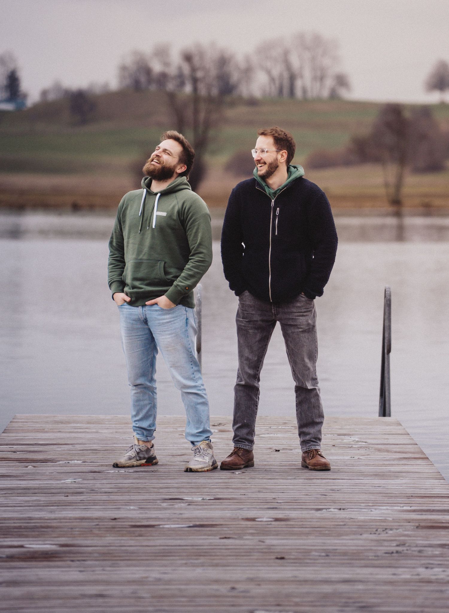 Two people stand together on a wooden dock overlooking a serene lake on an overcast day.