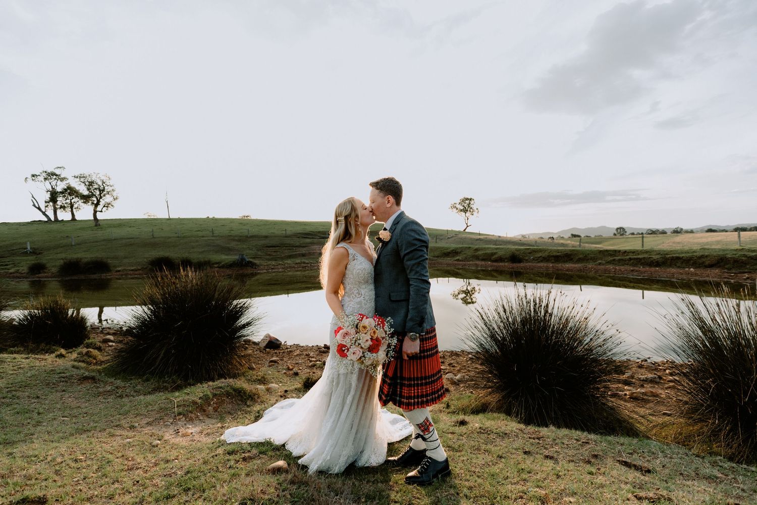 Romantic wedding portrait of a bride and groom kissing by a lake at sunset, showcasing natural light photography.