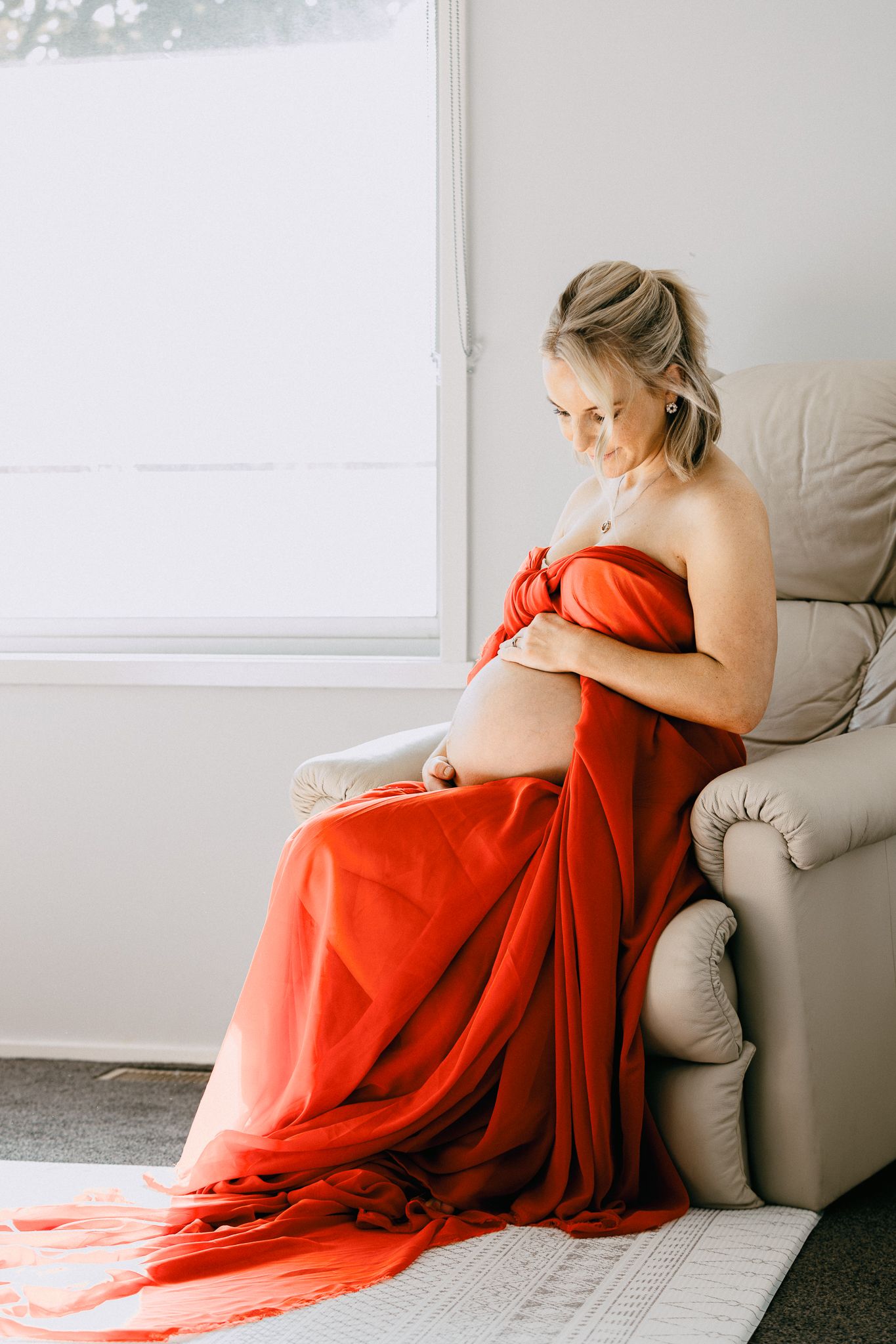 Maternity portrait of a woman wrapped in a flowing red fabric sitting in a chair by a bright window.