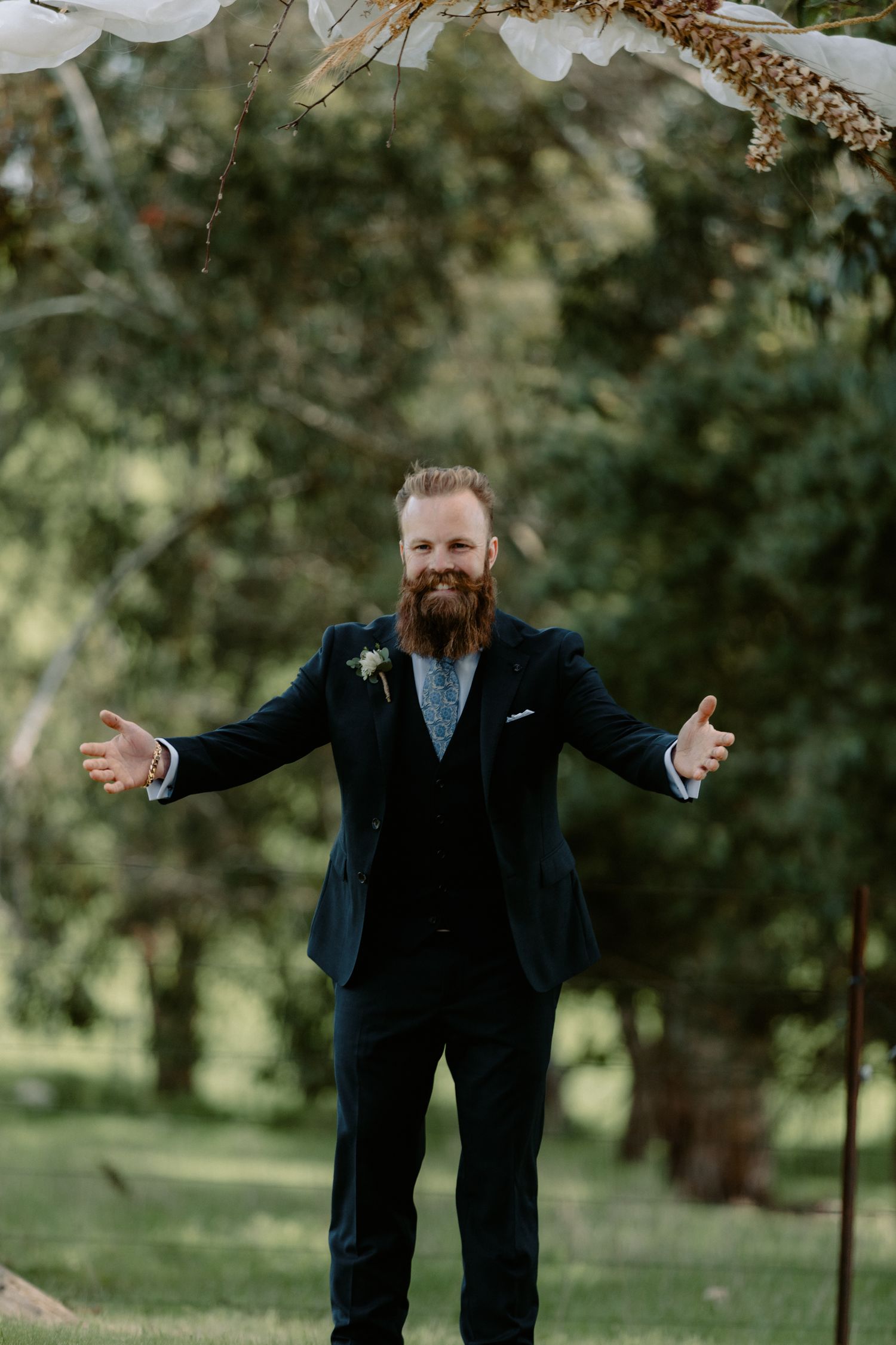 Groom with a large groomed beard and navy suit waiting at the altar with arms wide open during an outdoor Melbourne wedding.