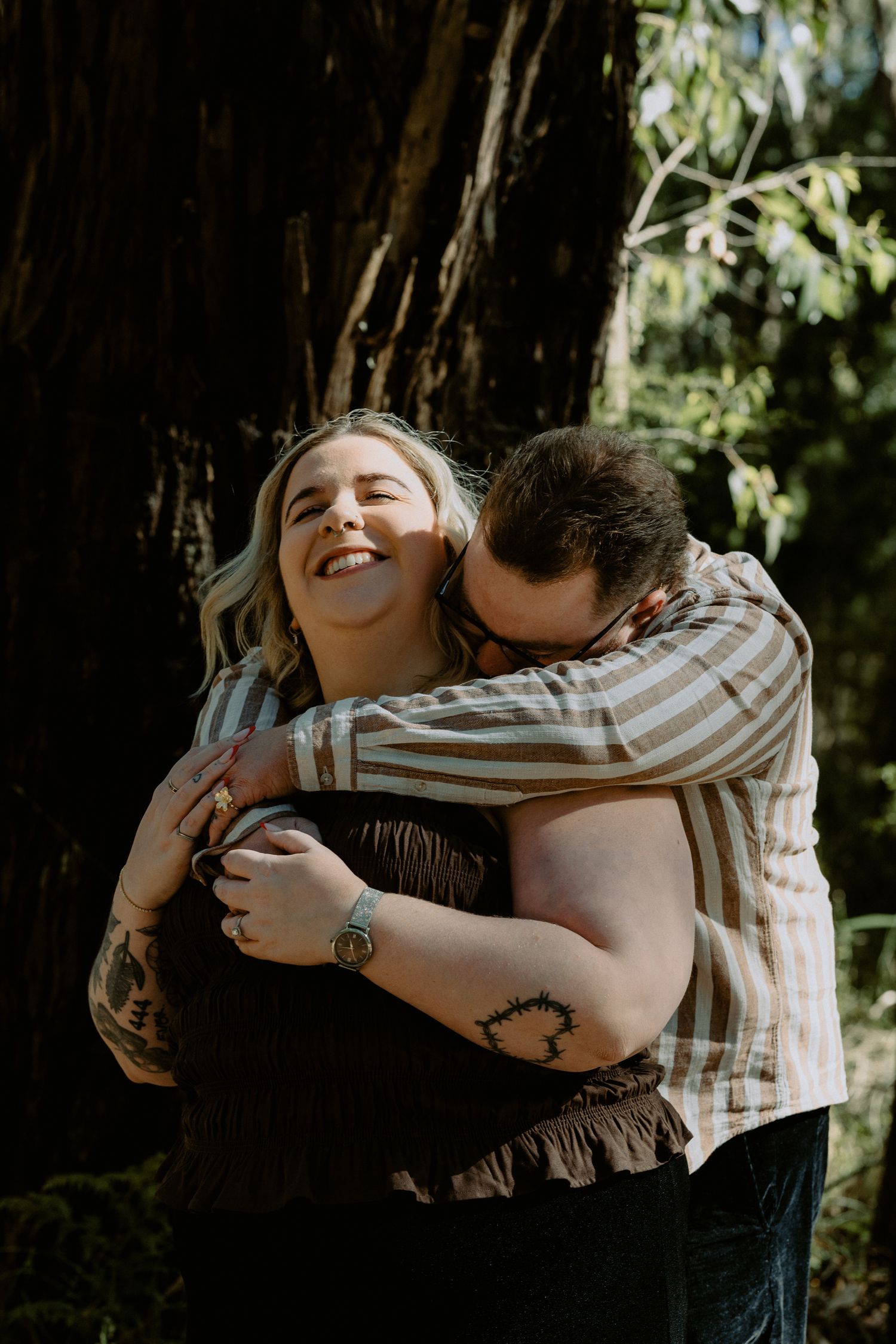 Joyful newly engaged couple embracing during a forest photoshoot, capturing candid laughter and a rustic adventure aesthetic.