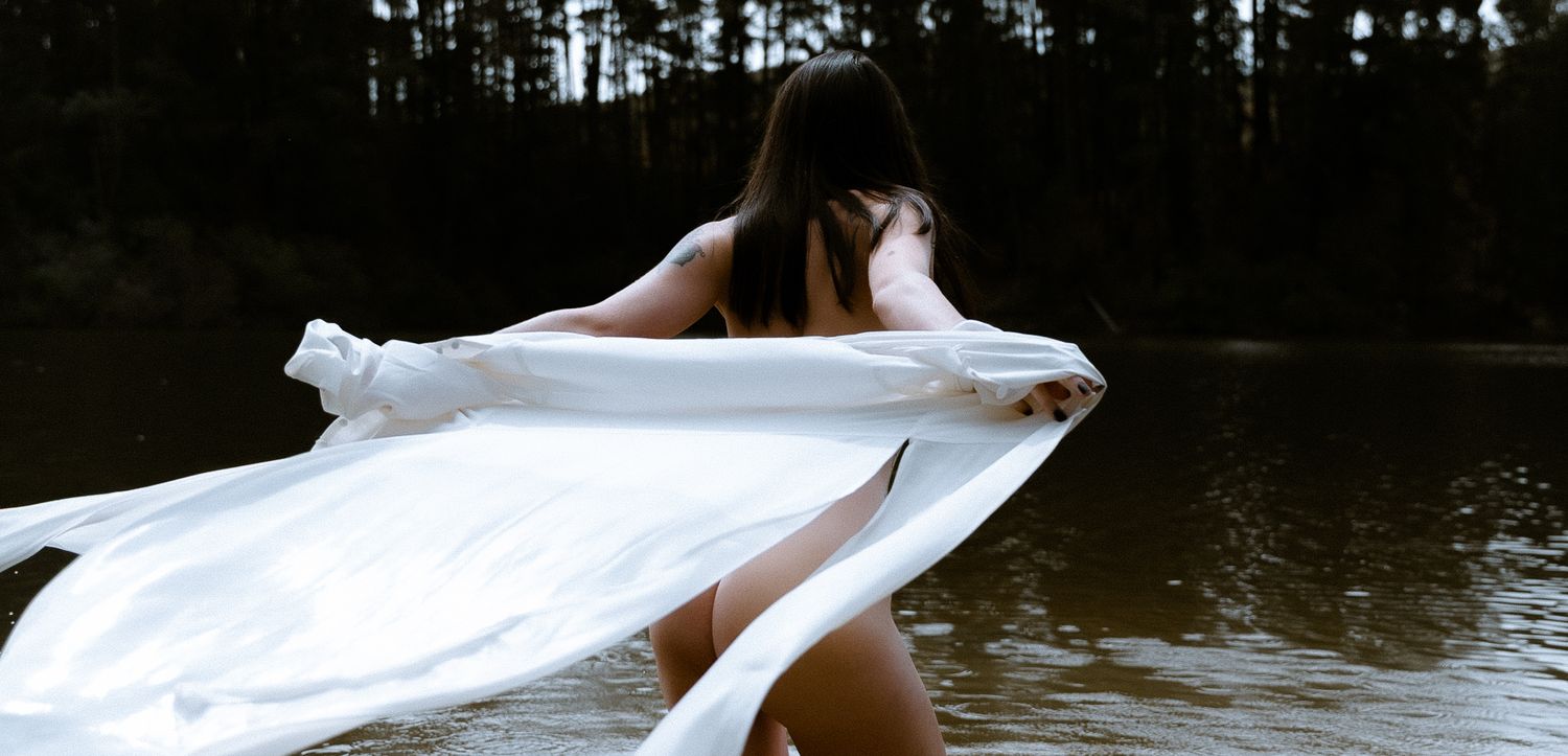 Artistic lifestyle photography of a woman walking into a lake at dusk, draped in a flowing white shirt for a moody editorial session.