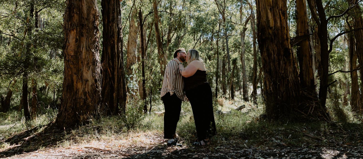 Romantic forest engagement photoshoot of a couple kissing between towering gum trees in a natural bushland setting.