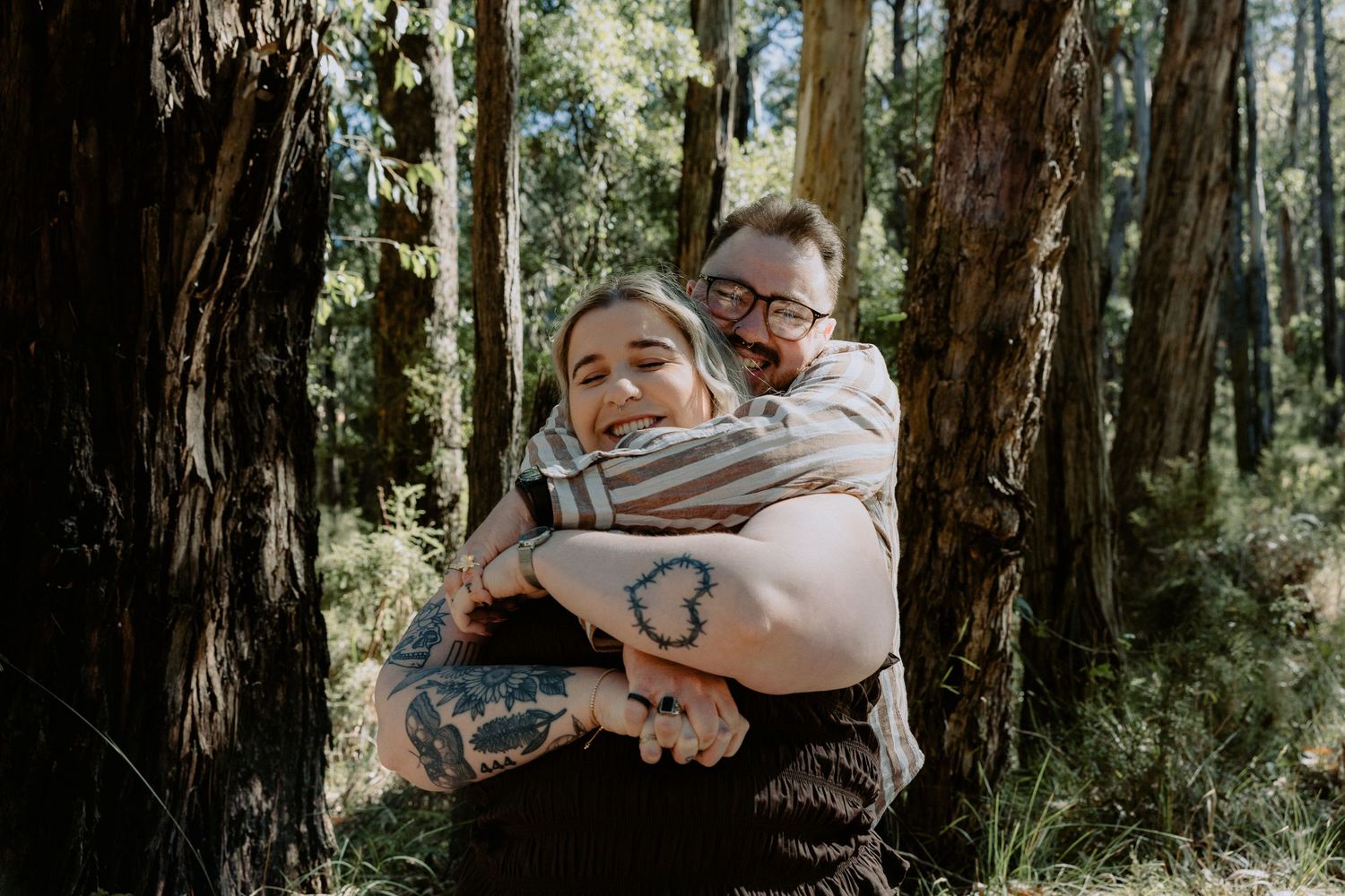 Joyful newly engaged couple embracing during a forest photoshoot, capturing candid laughter and a rustic adventure aesthetic.