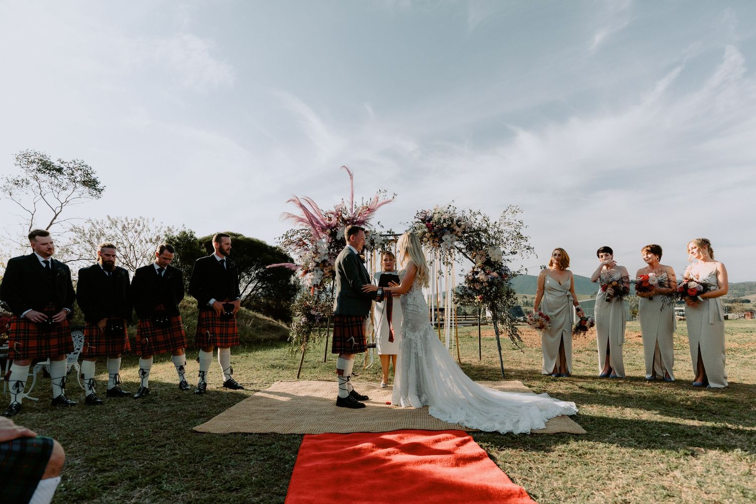 Candid outdoor wedding ceremony featuring a bride in a lace gown and a groom in a kilt at a scenic farm venue.