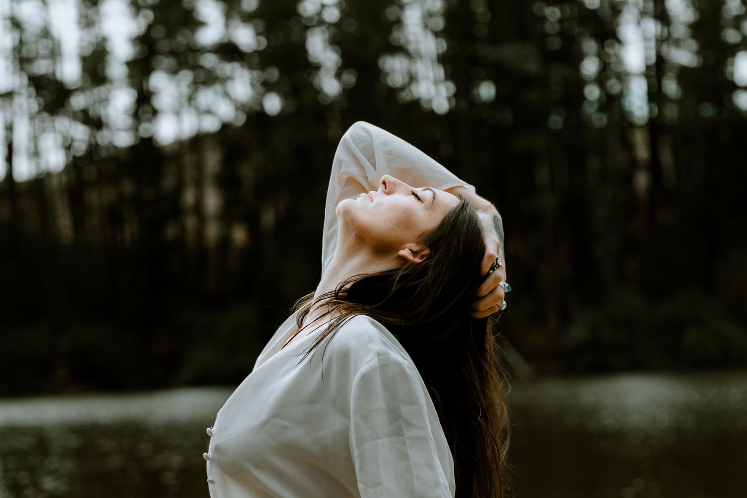 Moody outdoor portrait of a woman in a white silk blouse with eyes closed, leaning back in a tranquil forest and lake setting.