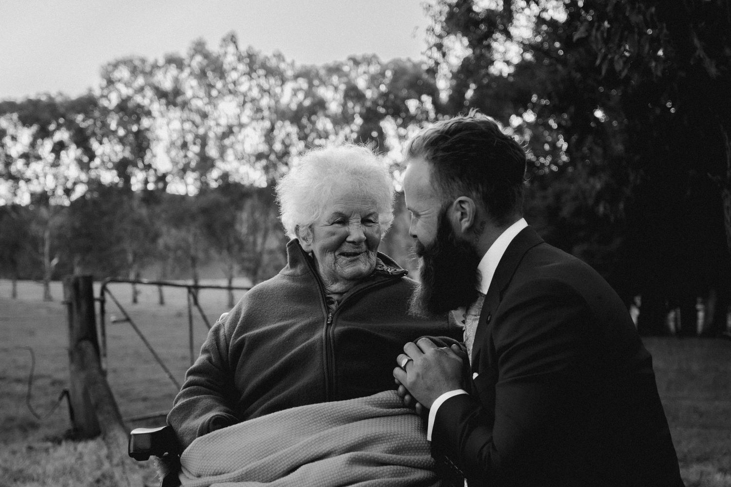 Black and white documentary-style wedding photography of a groom sharing a heartfelt moment with his grandmother.