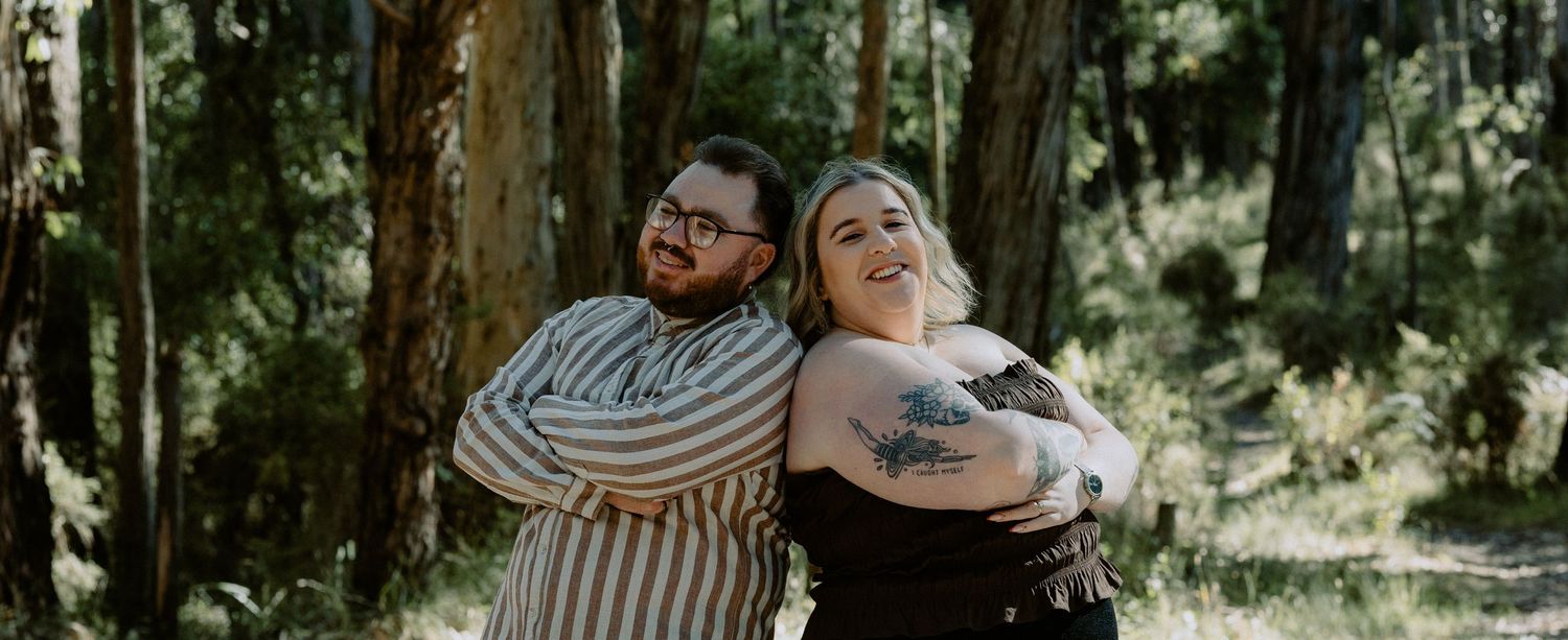 Fun engagement portrait of a couple standing back-to-back with arms crossed in a sunny woodland setting.