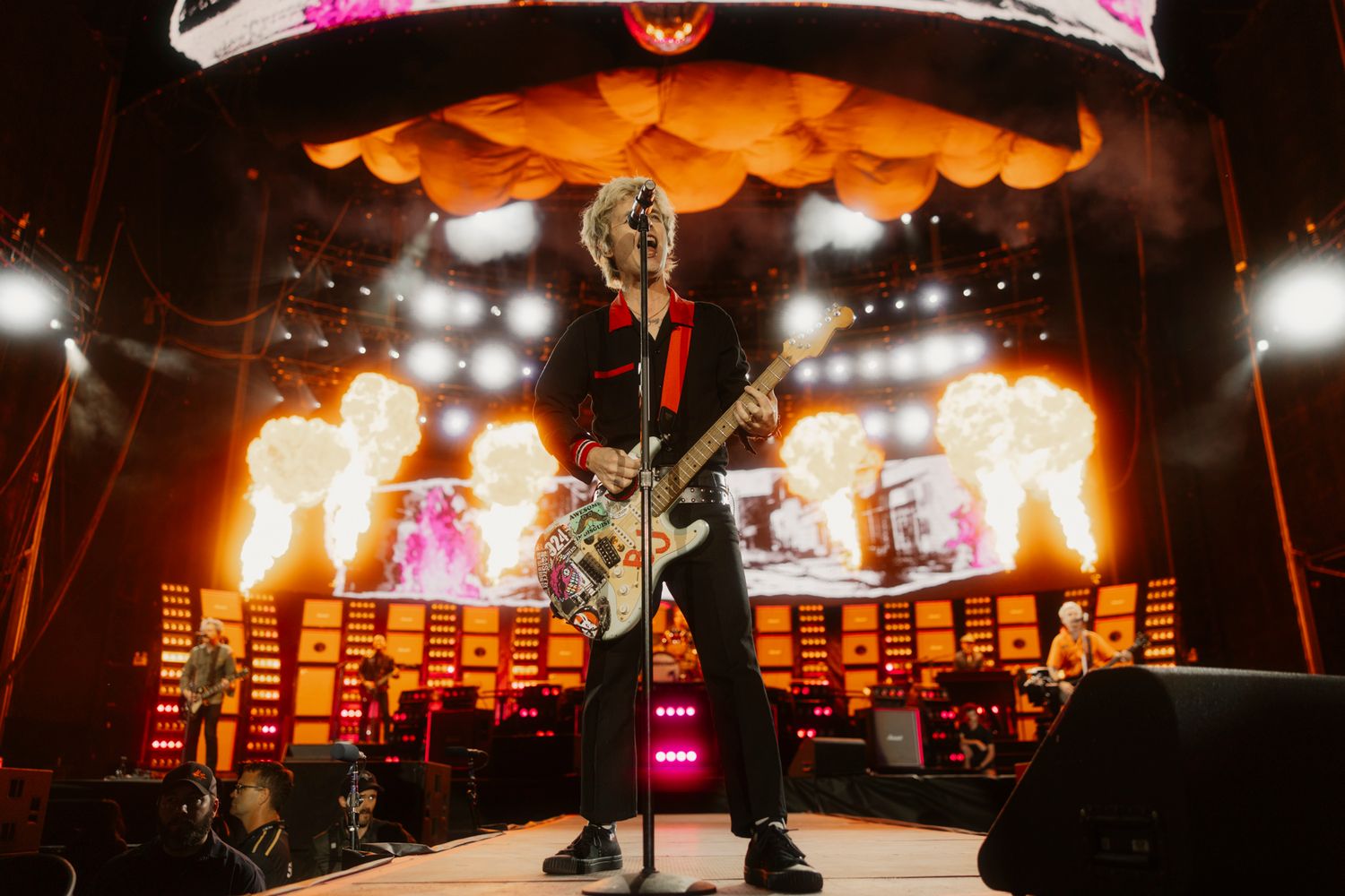 Green Day live on the catwalk with pyrotechnic flames. Wide-angle professional concert photography capturing a stadium rock show.