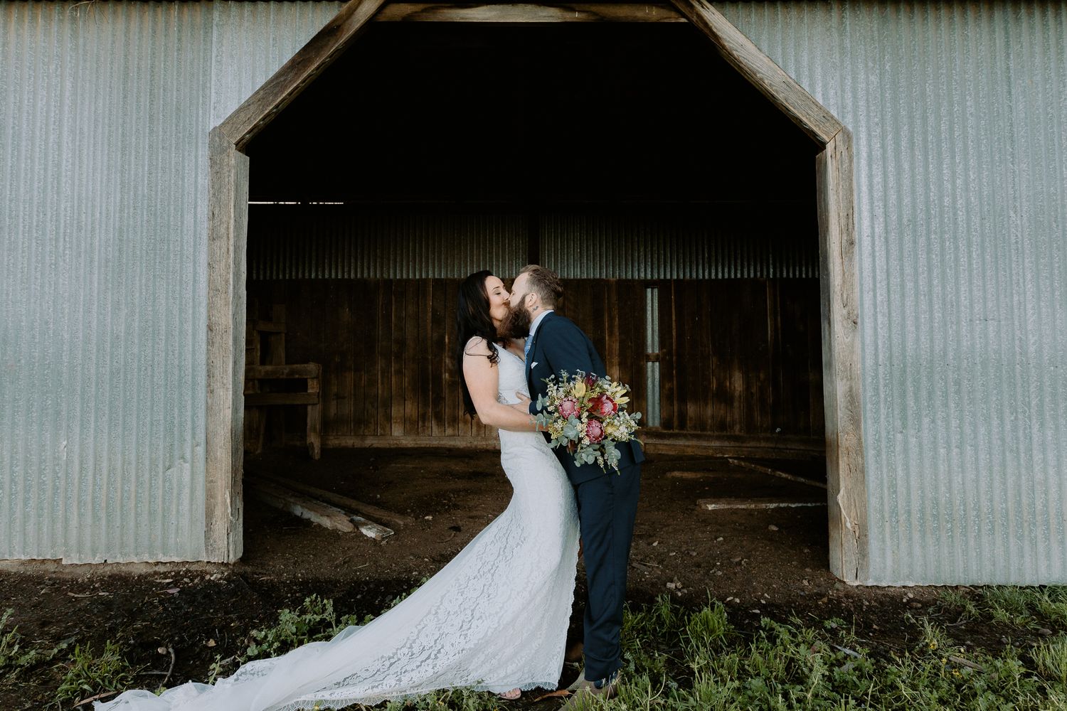 Romantic kiss between a bride and groom in the doorway of a large rustic barn on a farm property.
