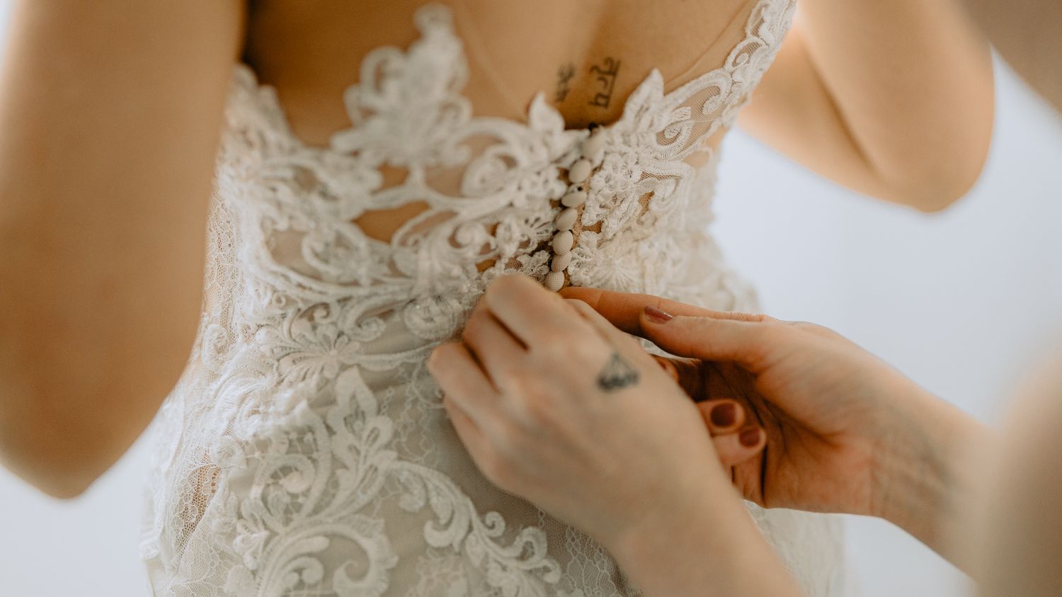 Close-up of a bride's lace wedding dress being buttoned up, showing intricate floral embroidery and a small hand tattoo.