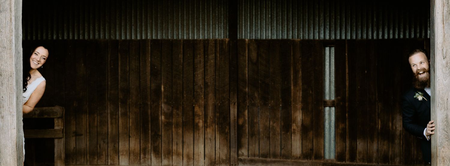 Playful wedding portrait of a bride and groom peeking out from behind the large wooden doors of a rustic barn.
