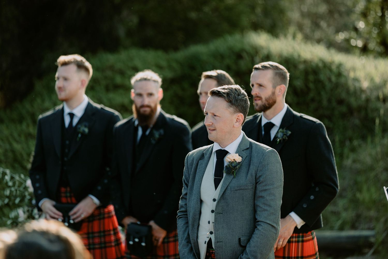 Groom and groomsmen in traditional kilts waiting at the altar during a rustic outdoor wedding ceremony.