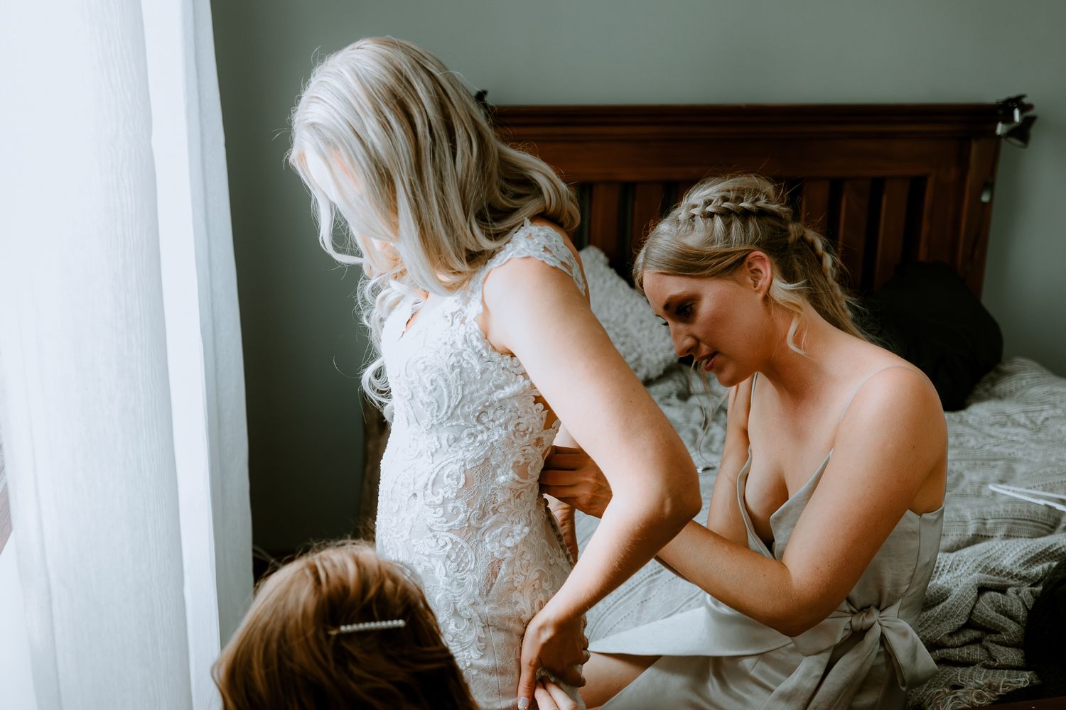 Behind the scenes of a bride getting ready with bridesmaids helping her into a white lace wedding gown by a window.