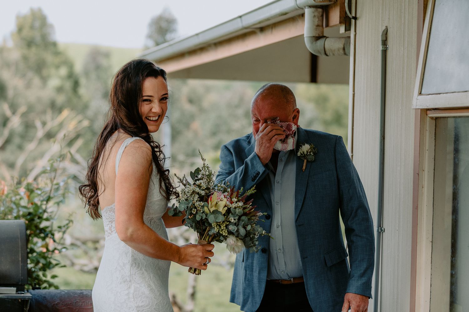 Candid wedding photography of a laughing bride in a lace gown next to her father wiping away tears of joy.
