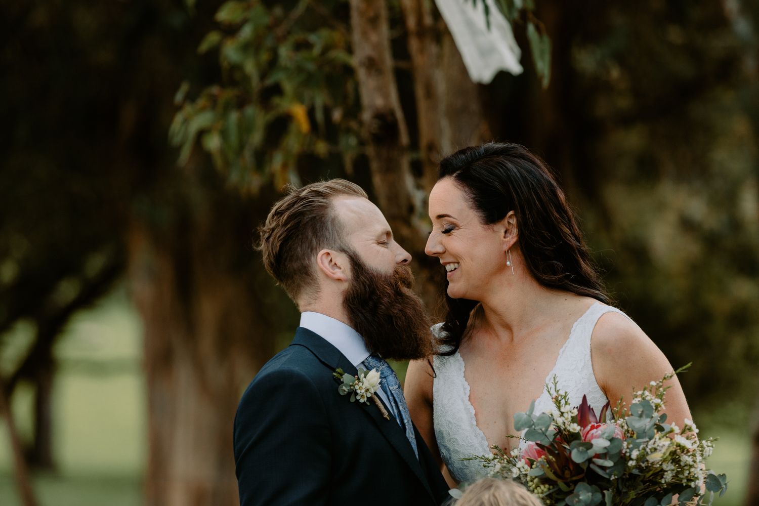Close-up romantic portrait of a bride and a groom with a large beard smiling at each other during an outdoor ceremony.