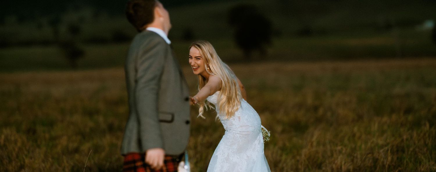 Candid wedding photography of a laughing bride in a lace wedding dress and groom in a traditional Scottish kilt during a sunset photoshoot.